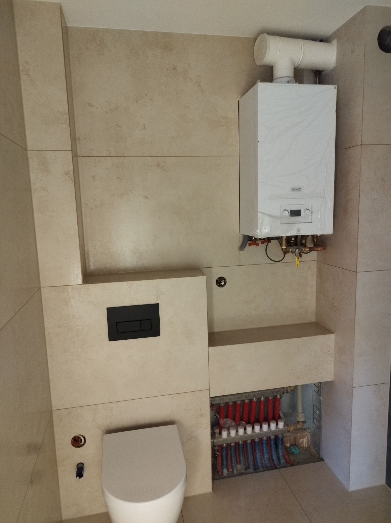 Beżowa łazienka z podtynkową toaletą, grzejnikiem w folii i rozdzielaczem ogrzewania podłogowego. Minimalistyczny design z dużymi płytkami.