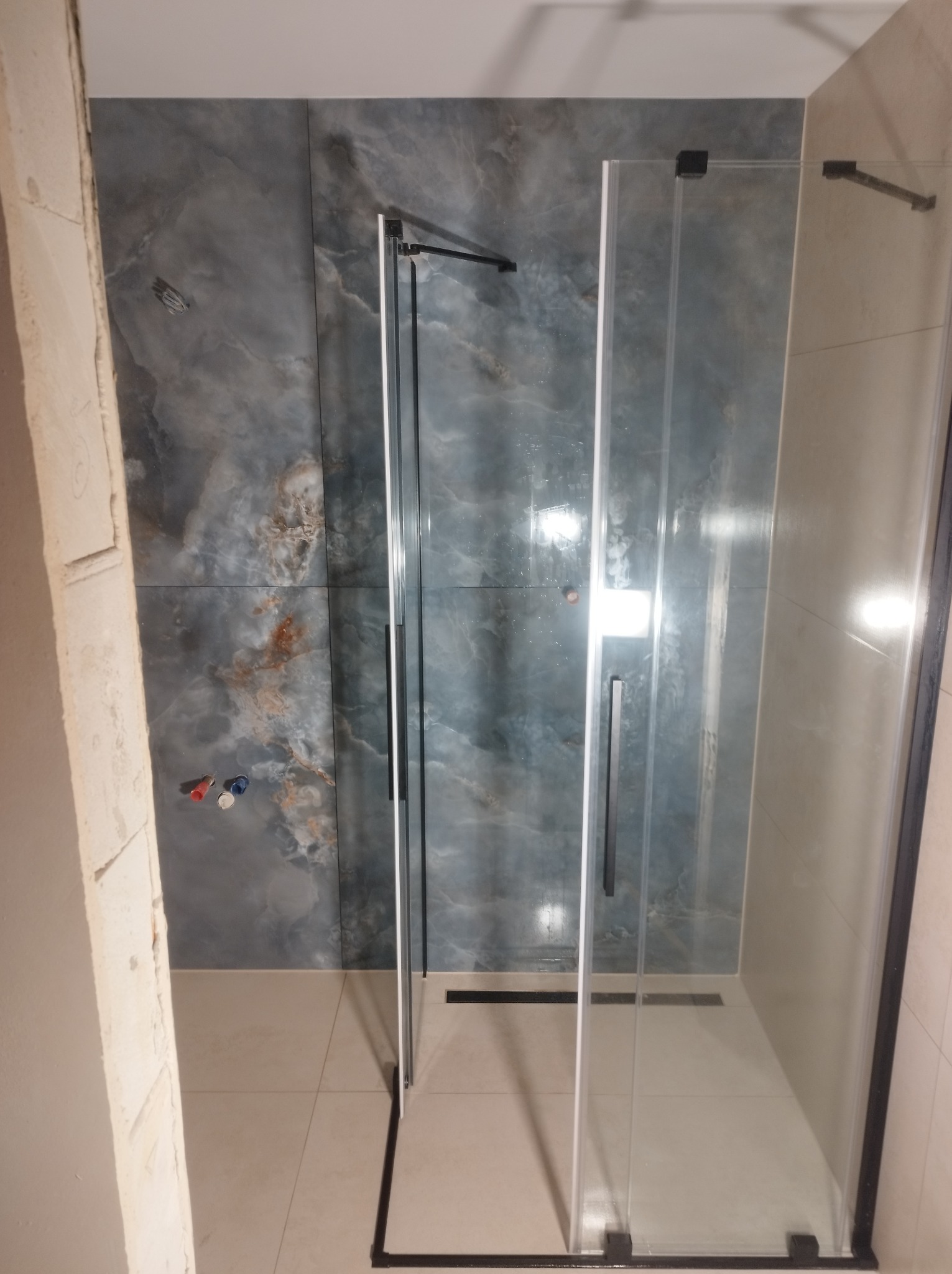 Nowoczesna kabina prysznicowa z czarnymi profilami i marmurowymi płytkami w odcieniach szarości i beżu, widok z góry, fragment surowej ściany po lewej.