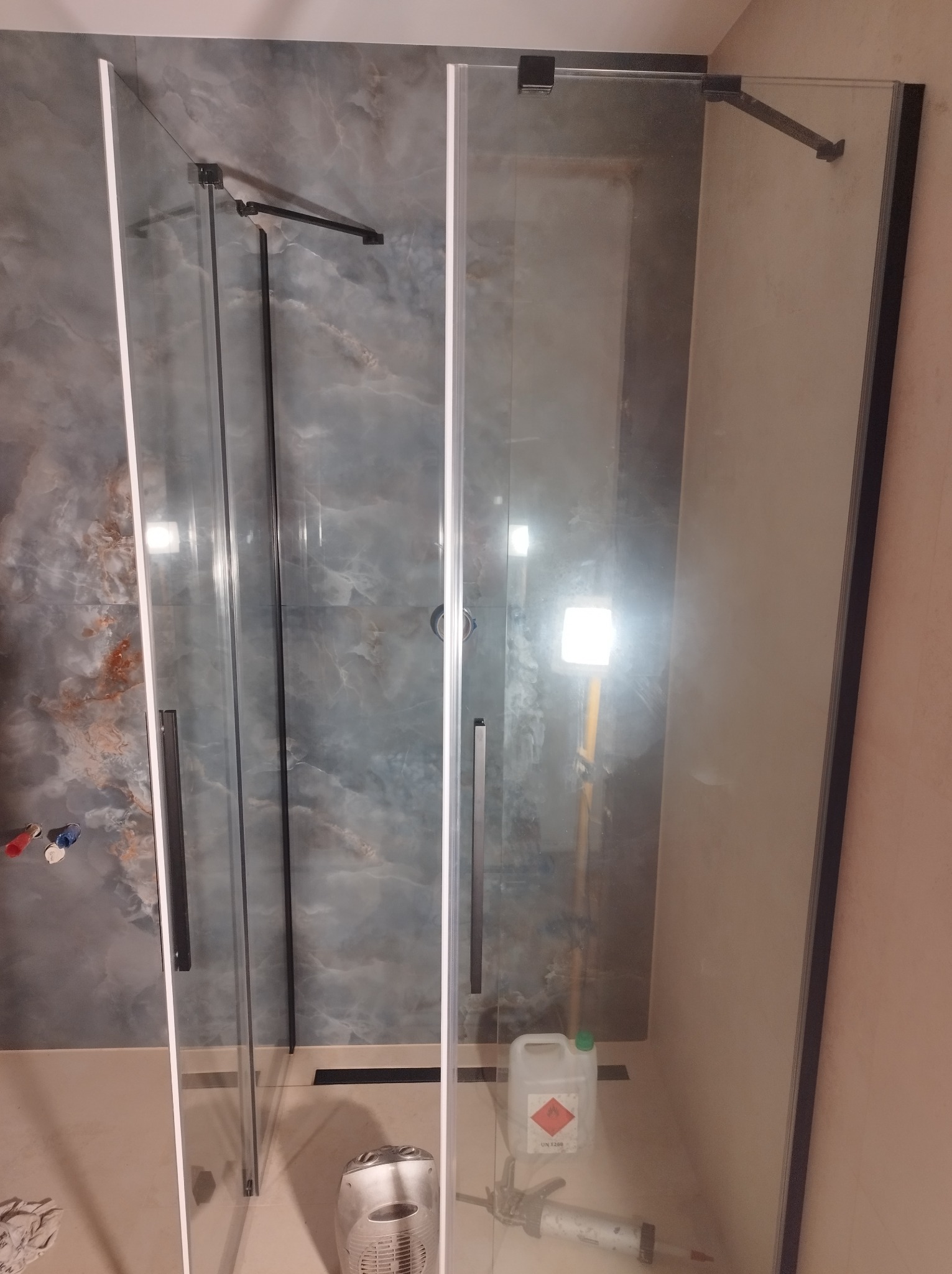 Nowoczesna kabina prysznicowa z czarnymi detalami na tle marmurowej ściany. Widoczne narzędzia i chemia budowlana sugerują prace wykończeniowe.