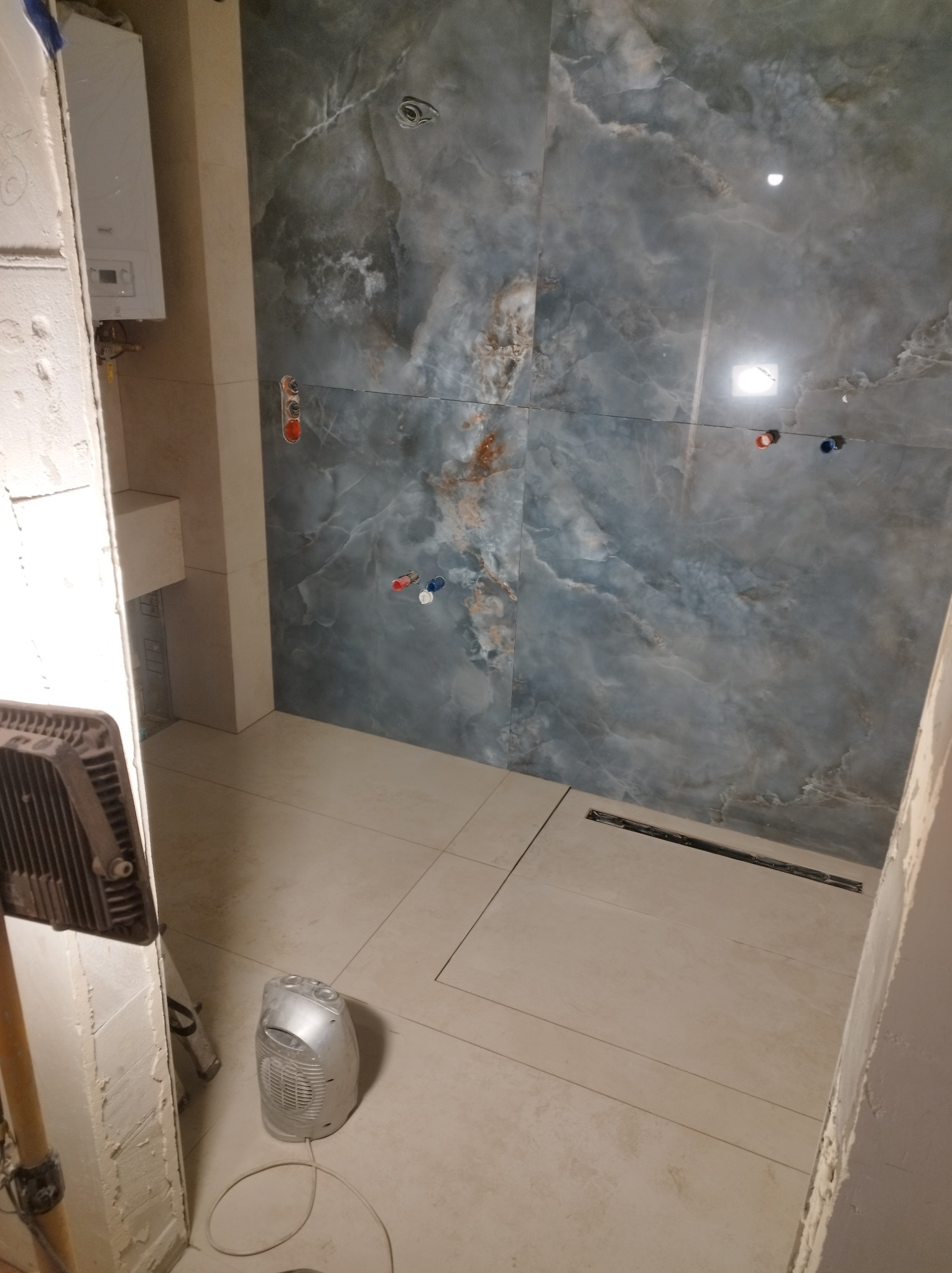 Łazienka w trakcie remontu: położone płytki imitujące marmur na ścianach i podłodze, widoczny odpływ liniowy, grzejnik elektryczny i lampa budowlana.