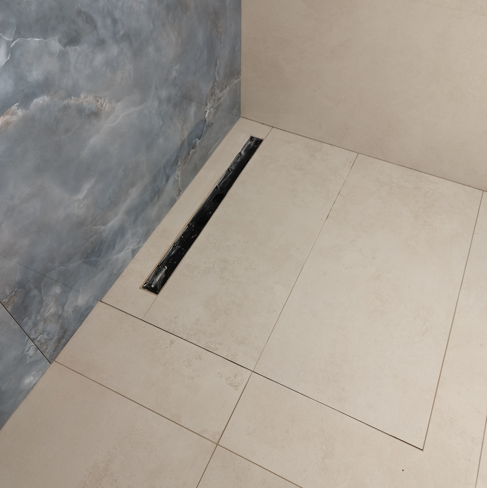 Beżowe płytki podłogowe w kabinie prysznicowej z odpływem liniowym i szarą okładziną ściany imitującą marmur. Minimalistyczny design.
