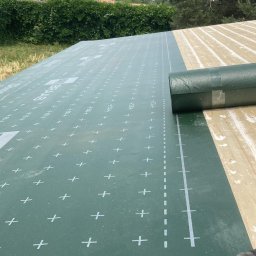 Dariusz Prejna - Zielona membrana dachowa z nadrukiem układana na drewnianej konstrukcji dachu, w tle widok na zielone wzgórza i zabudowania.