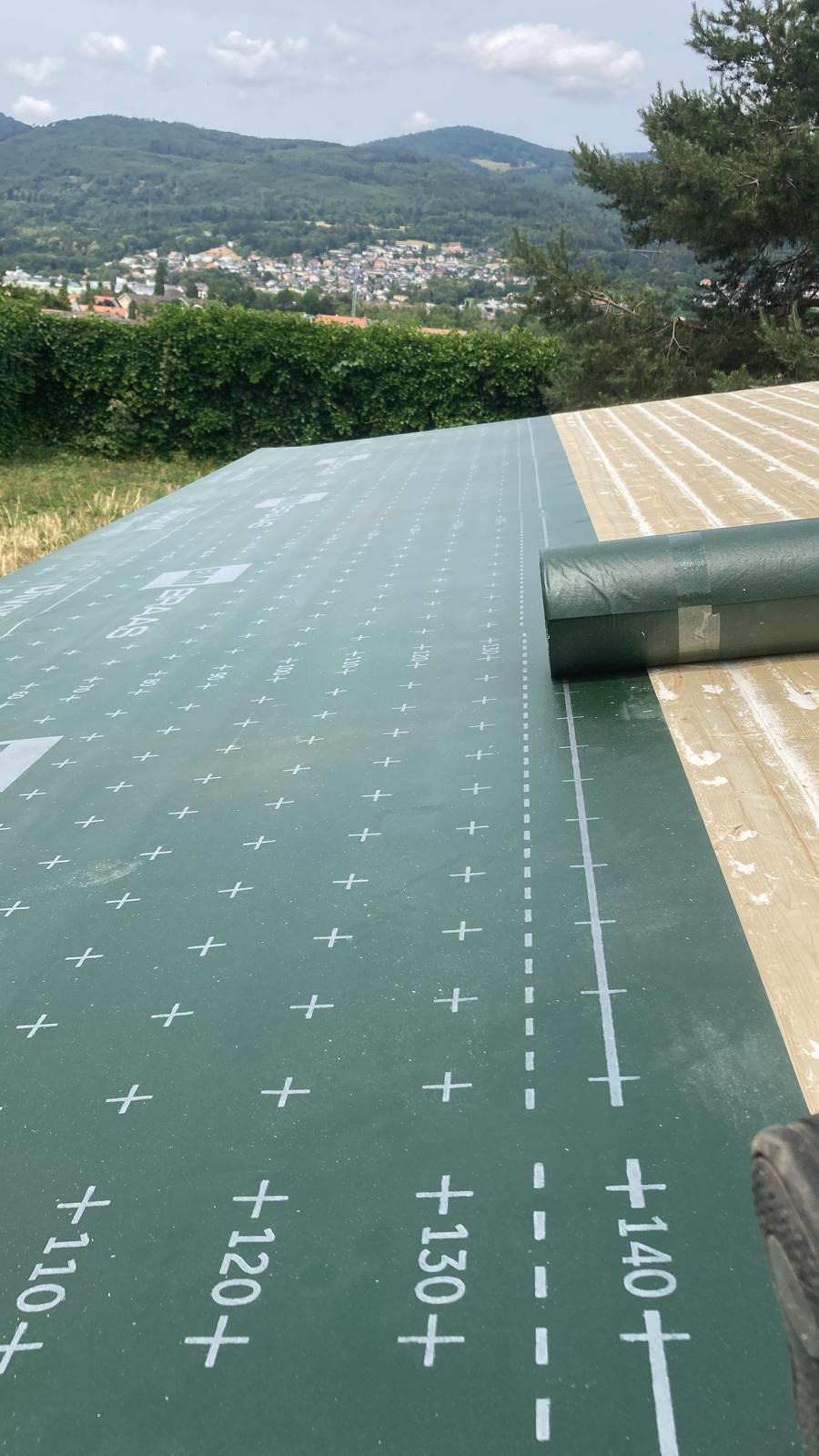 Zielona membrana dachowa z nadrukiem układana na drewnianej konstrukcji dachu, w tle widok na zielone wzgórza i zabudowania.