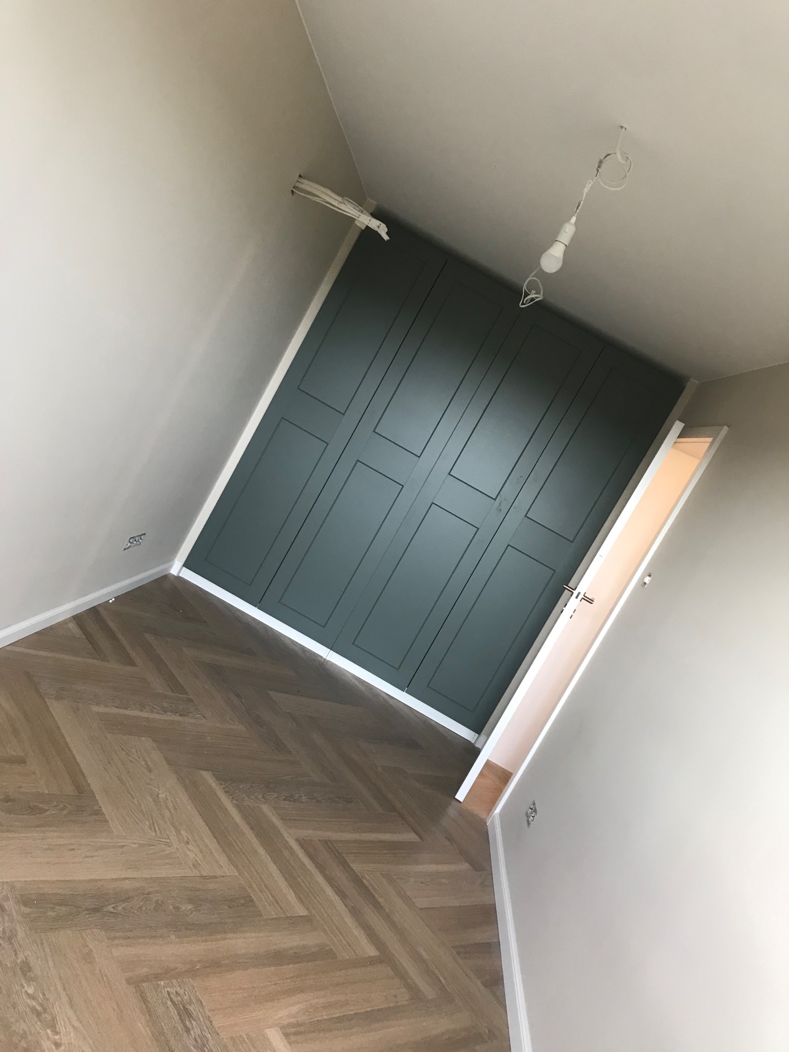 Pokój po remoncie: parkiet w jodełkę, ściany w neutralnych barwach, zabudowa szafy w kolorze butelkowej zieleni z dekoracyjnymi panelami. Widoczne niedokończone oświetlenie.
