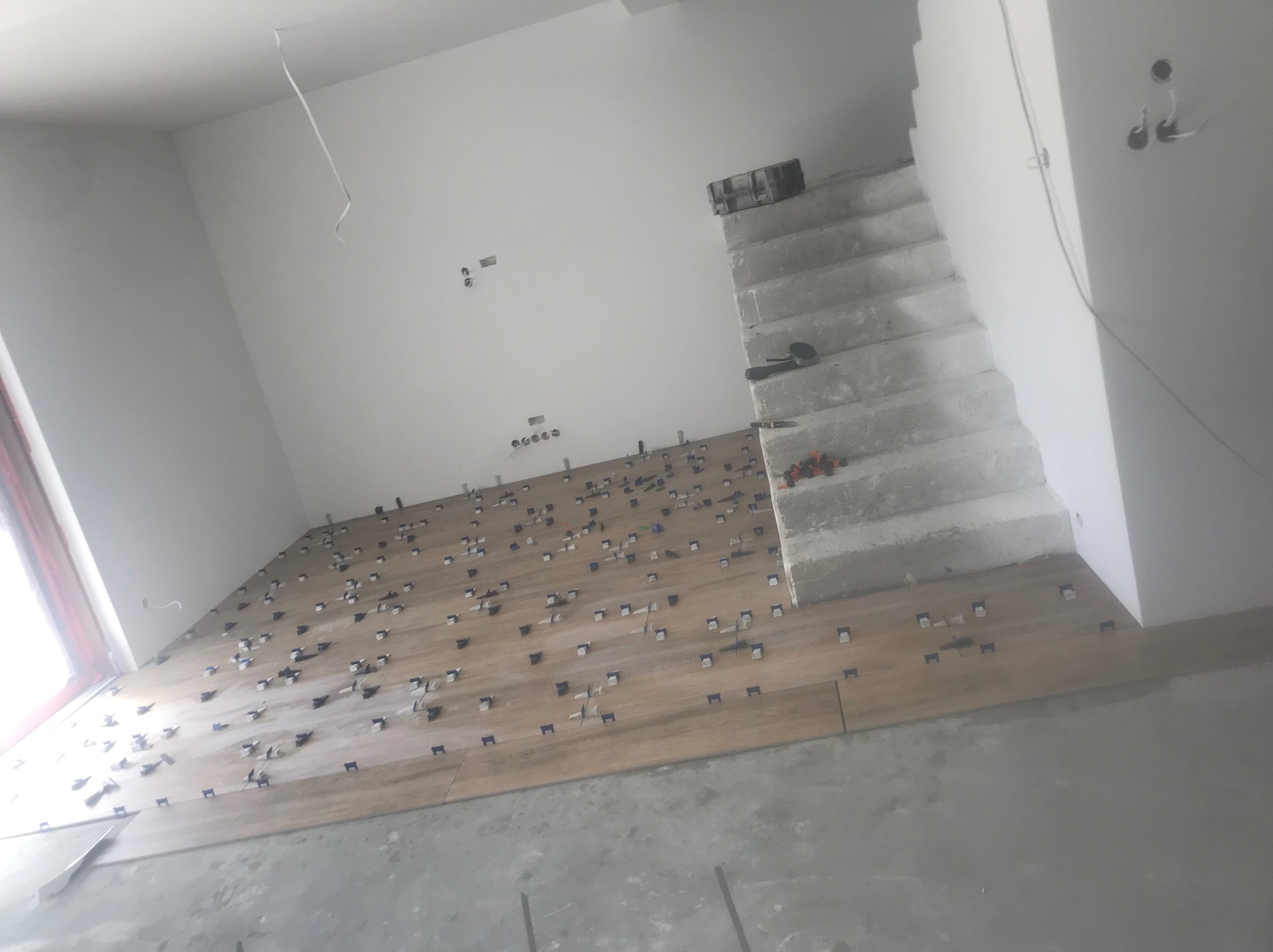 Układanie płytek imitujących drewno na podłodze w jasnym pomieszczeniu. Widoczne krzyżyki dystansowe i schody w tle. Prace glazurnicze w trakcie realizacji.