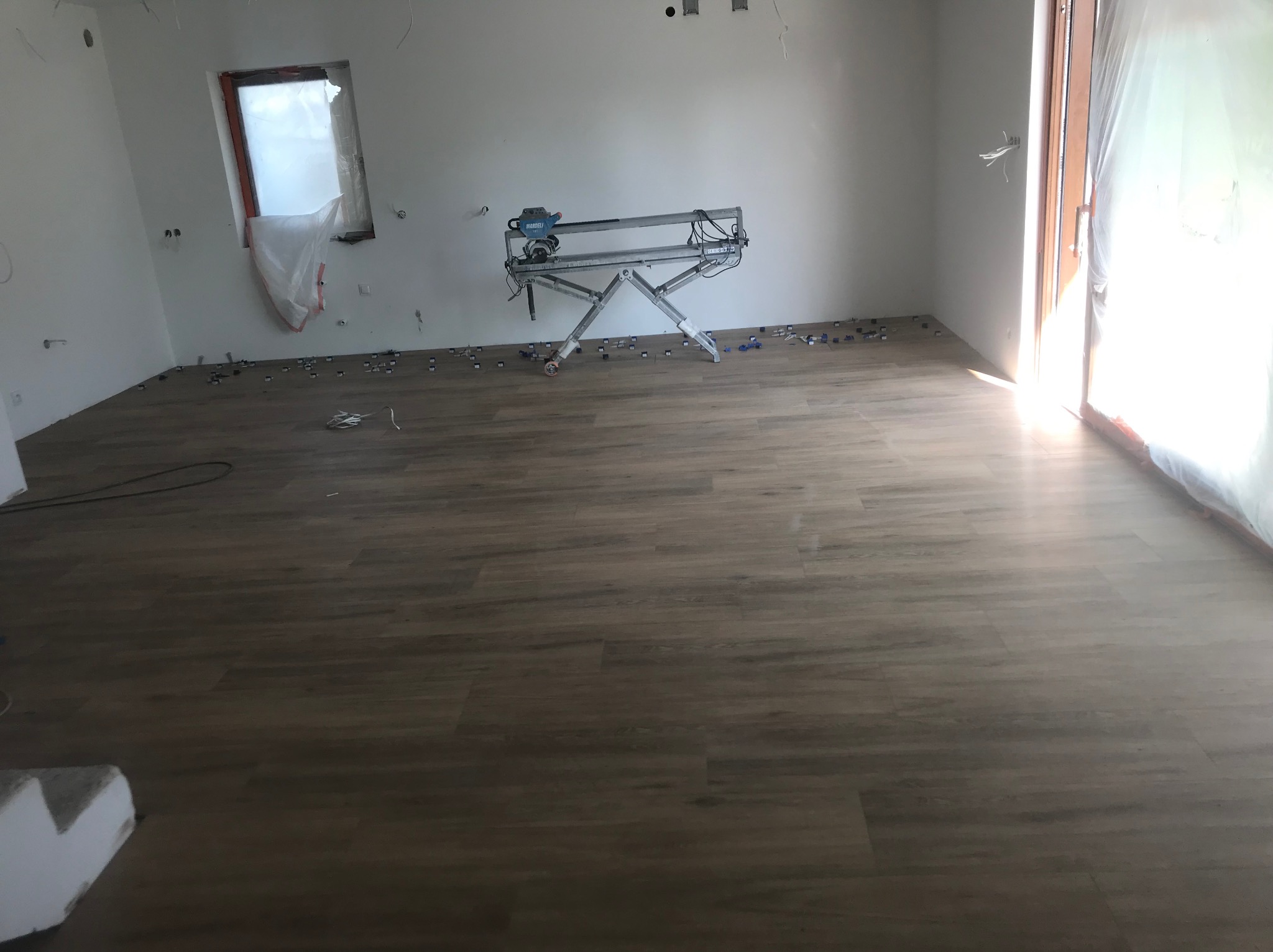 Ułożona podłoga z paneli w jasnym odcieniu drewna, z widoczną maszyną do cięcia paneli i elementami dystansowymi. Okno i drzwi zabezpieczone folią malarską.