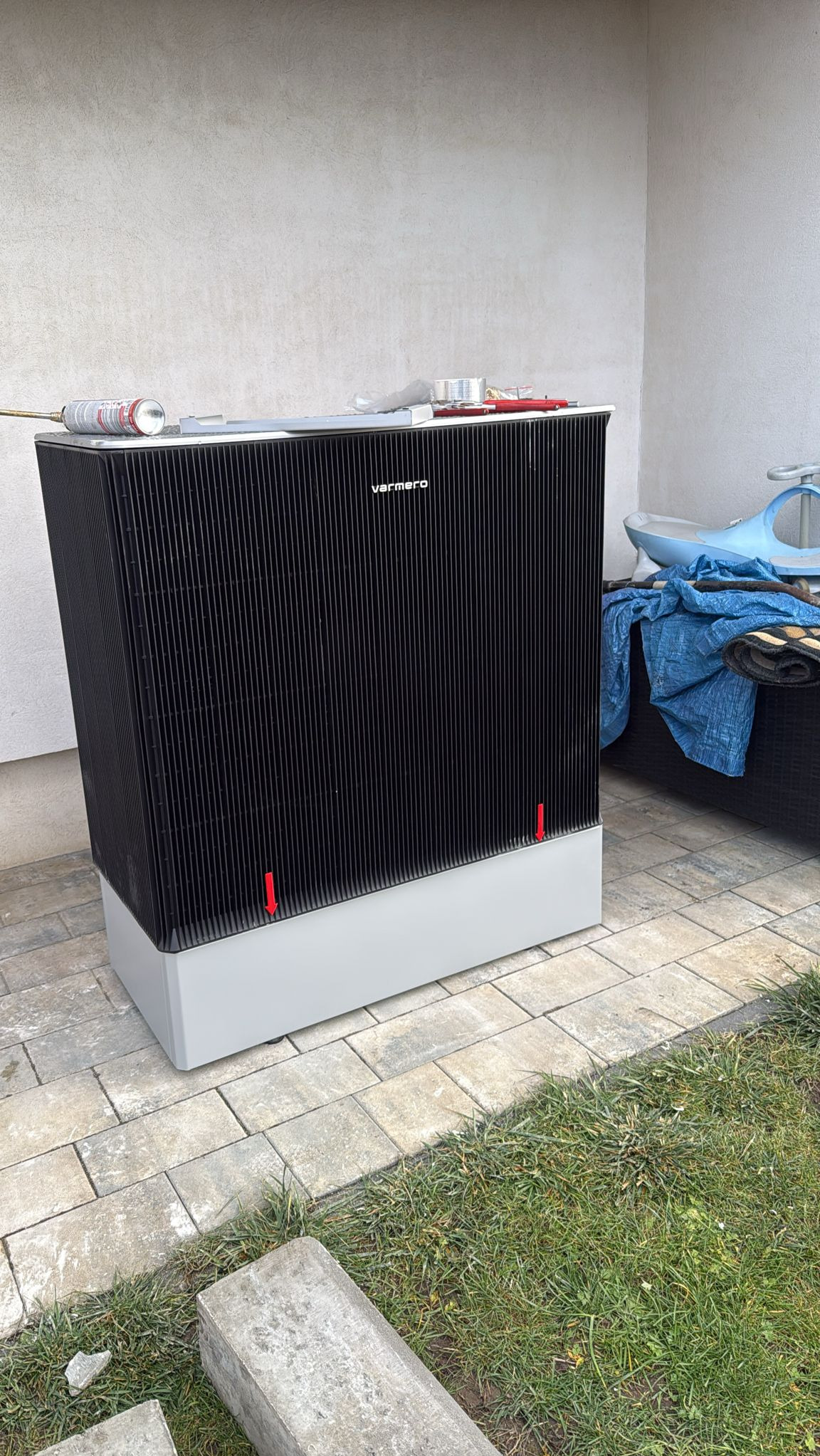 Czarna pompa ciepła Varmero na szarym cokole, z narzędziami na górze, stoi na kostce brukowej obok trawnika. Widok z boku, lekki kąt.