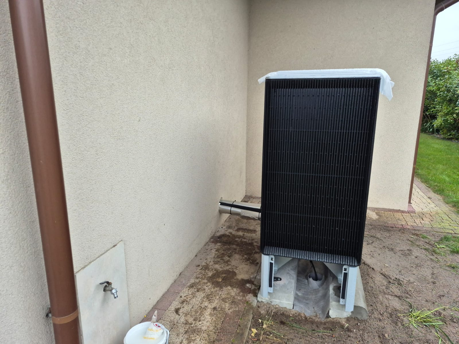 Zewnętrzna jednostka pompy ciepła powietrze-woda, zamontowana przy ścianie budynku z widocznymi rurami przyłączeniowymi i elementami montażowymi. Tło stanowi trawnik.