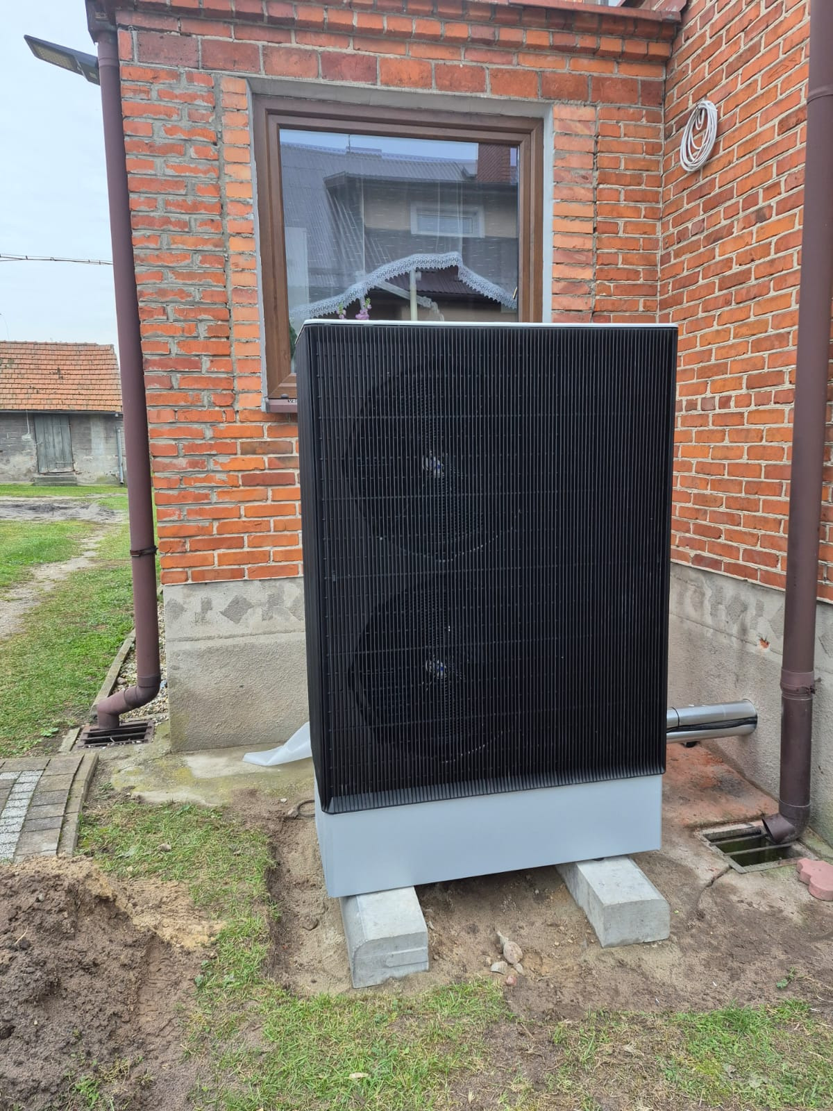 Czarna pompa ciepła powietrze-woda na betonowych blokach przy ceglanym budynku z brązową rynną. Widoczne okno z odbiciem dachu.