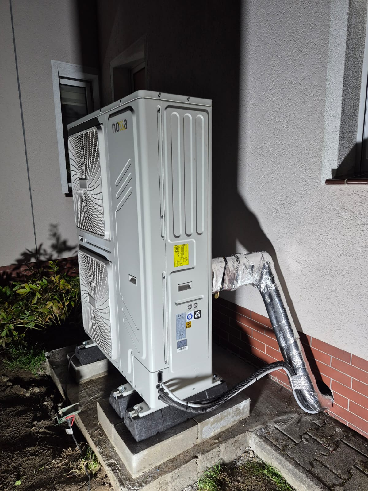 Biała, dwuwentylatorowa pompa ciepła marki Noxa, zamontowana na betonowej podstawie przy ścianie budynku z oknem. Rury izolowane folią aluminiową.