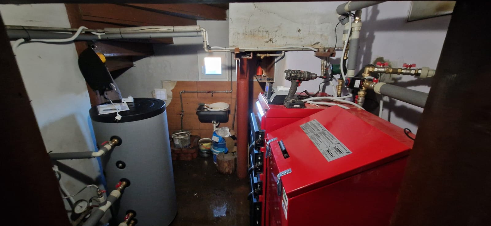 Czerwony piec grzewczy w piwnicy, obok szary zbiornik buforowy. Na piecu wiertarka akumulatorowa. Widoczne rury, zawory i instalacja. Małe okno w tle.