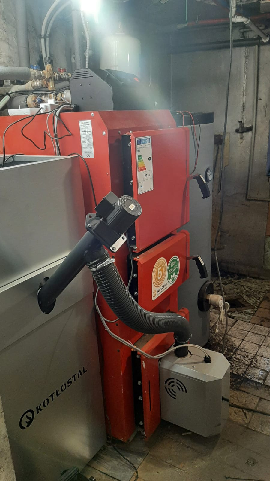 Czerwono-szary piec Kotłostal z widocznymi elementami instalacji, w kotłowni. Widoczne rury, przewody i etykiety informacyjne na urządzeniu.