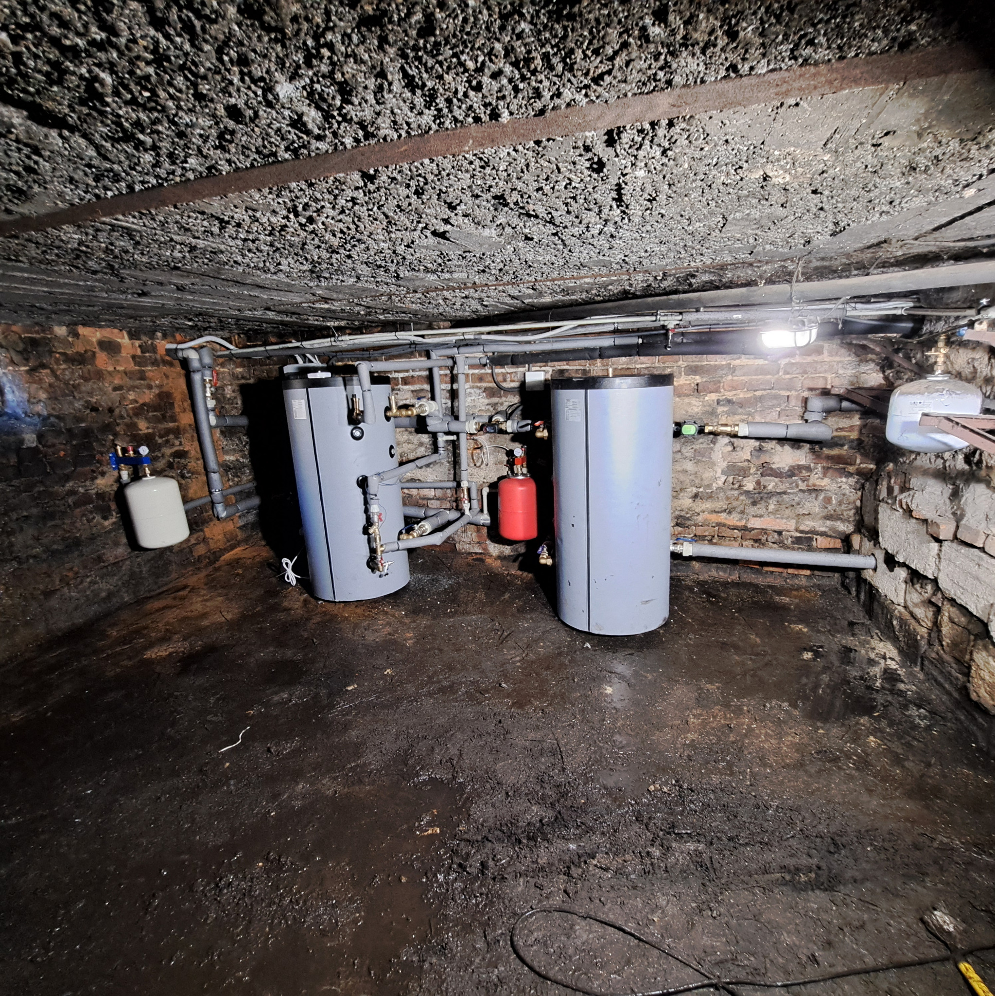 Dwa szare zbiorniki akumulacyjne ciepła w piwnicy z ceglanymi ścianami i betonowym sufitem. Instalacja hydrauliczna z rurami i zaworami. Widoczny mały, czerwony zbiornik.
