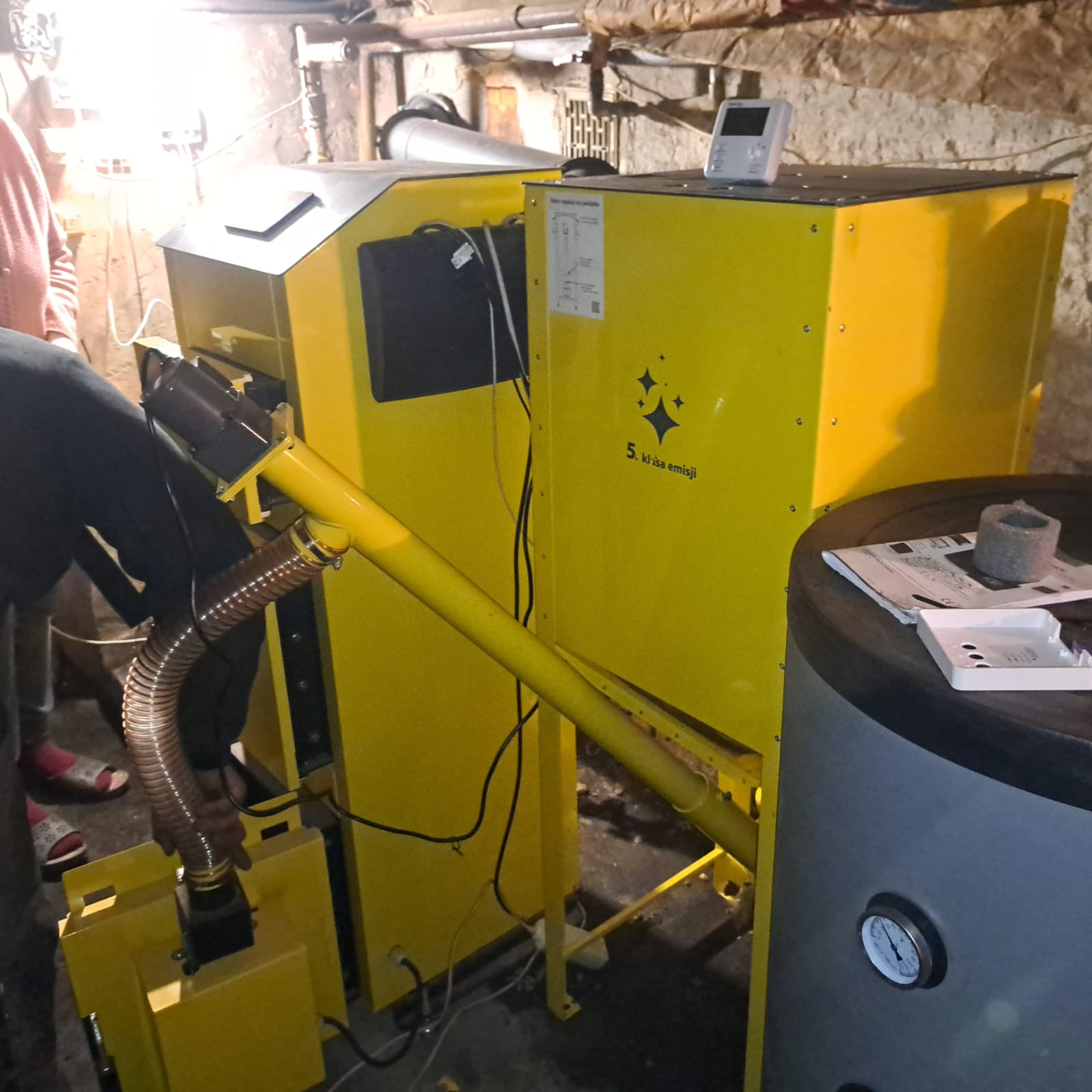 Żółty piec na paliwo stałe w kotłowni, widoczne elementy instalacji i podłączenia. Na piecu naklejka z klasą emisji. W tle ściana z rurami.