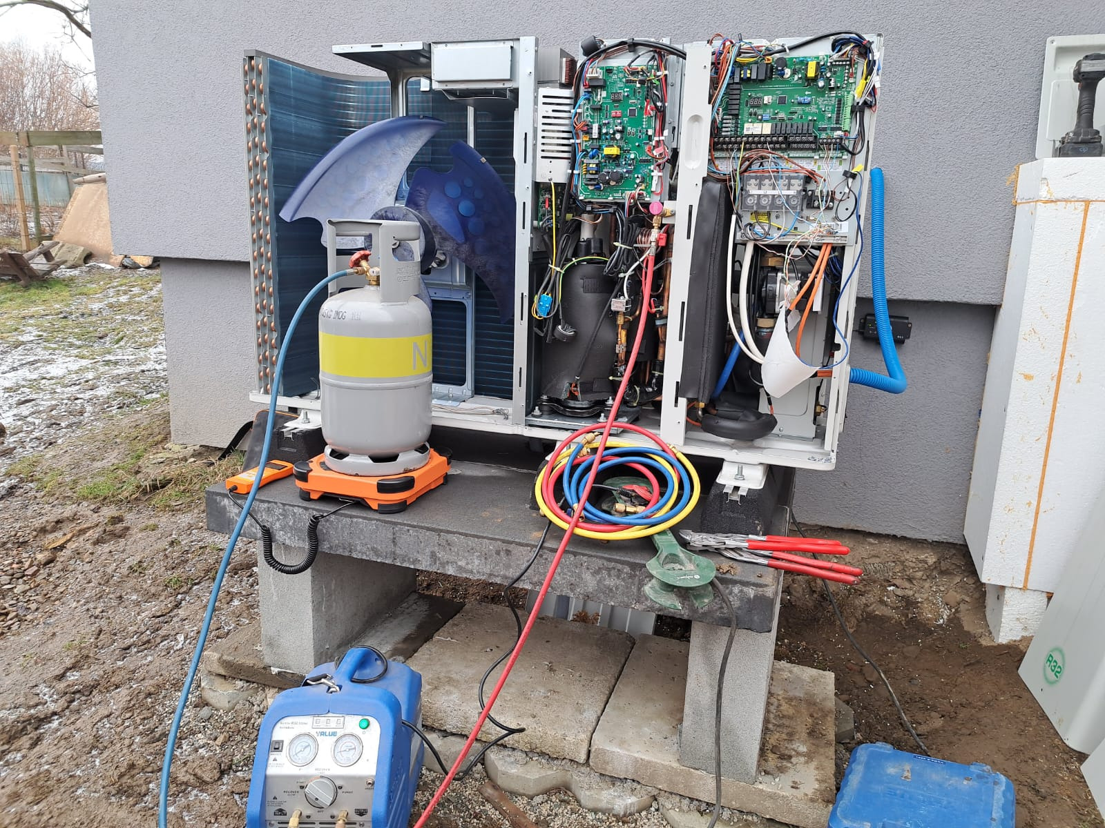 Otwarta obudowa pompy ciepła z widocznymi podzespołami, butla z gazem chłodniczym i narzędzia serwisowe na zewnątrz budynku. Przegląd instalacji.