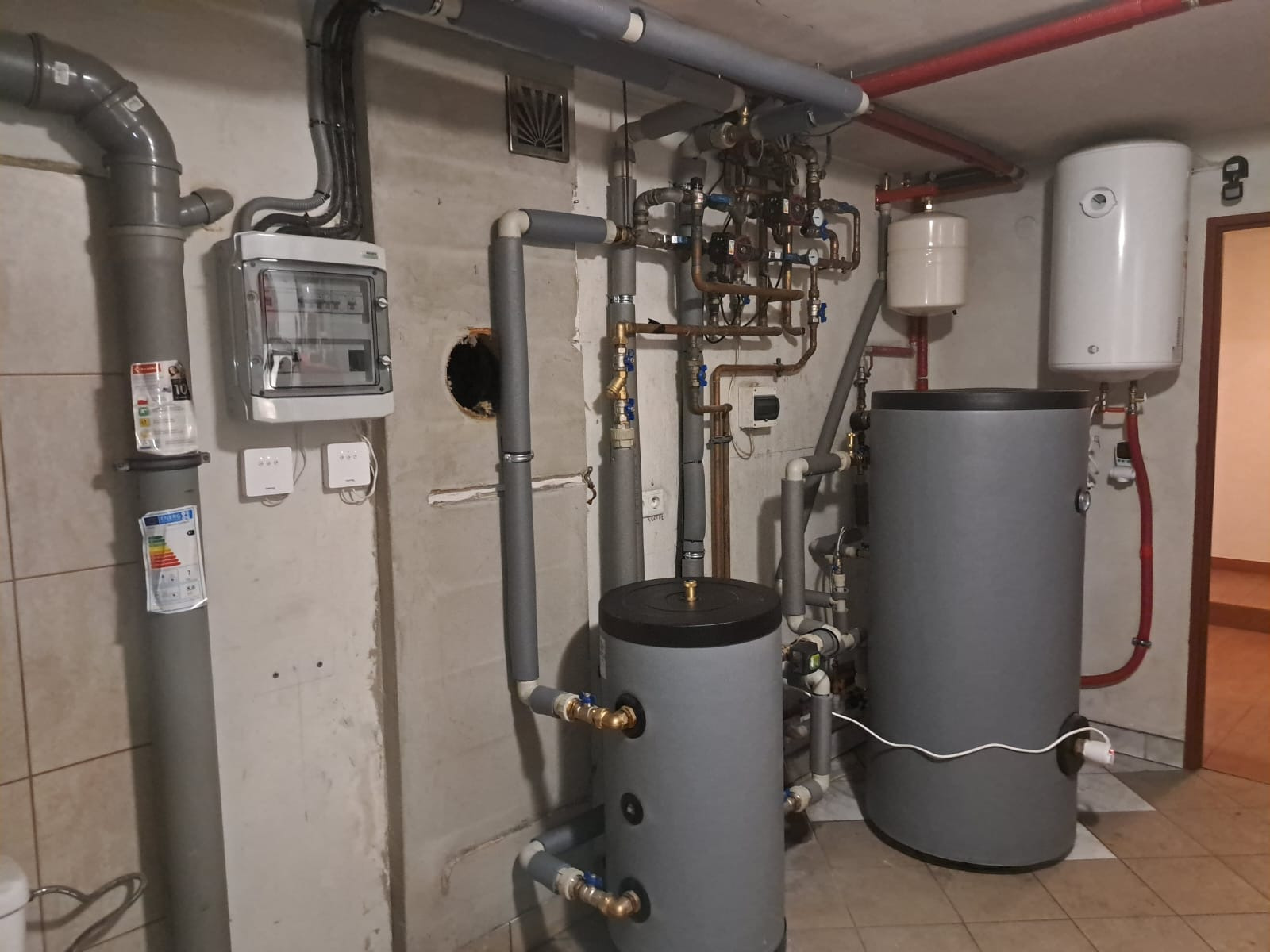 Piwnica z instalacją grzewczą: dwa szare zbiorniki, rury miedziane i izolowane, rozdzielnica elektryczna na ścianie. Widoczne elementy instalacji hydraulicznej.