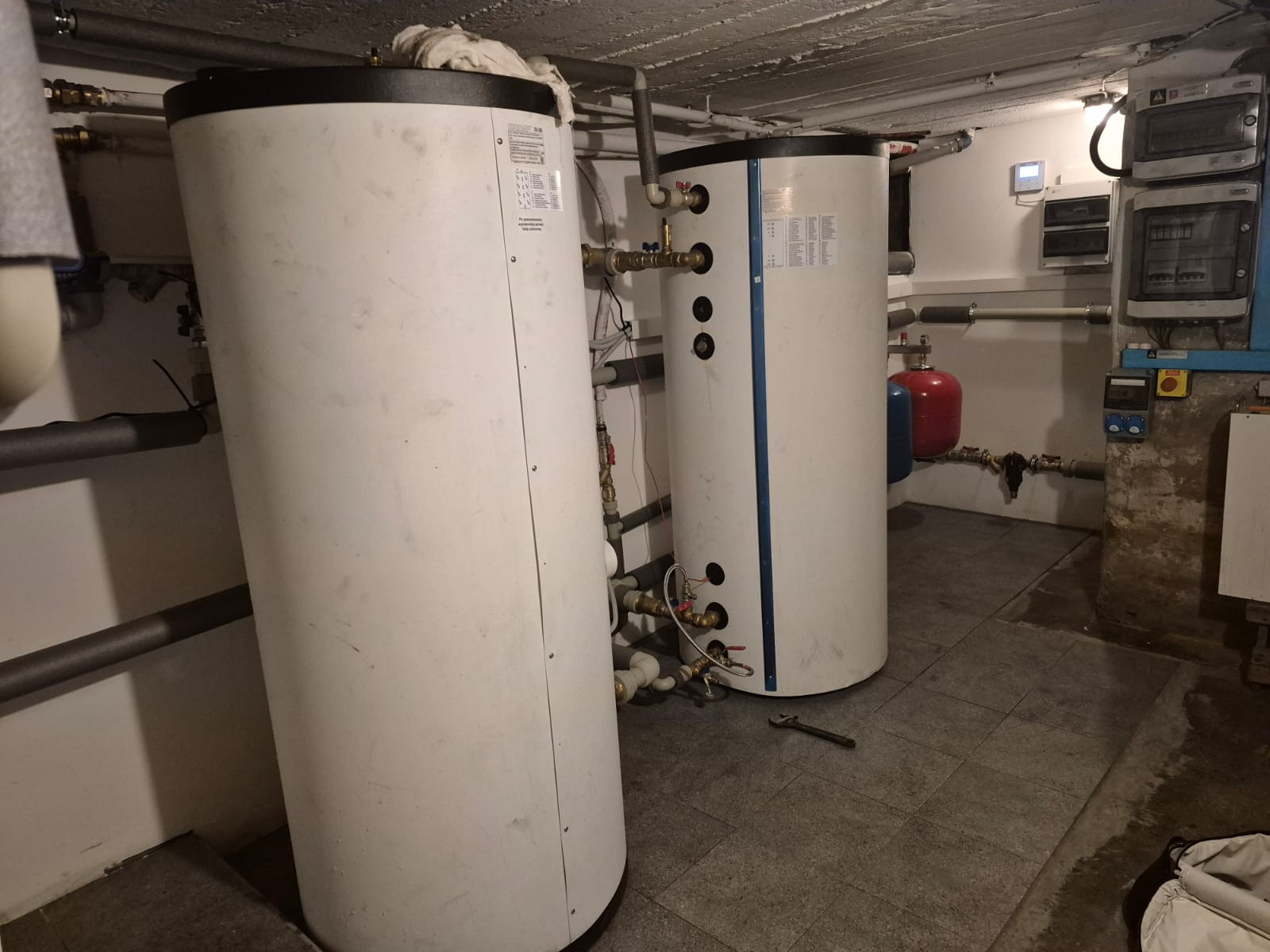 Dwa białe, cylindryczne zbiorniki z instalacją hydrauliczną w piwnicy, widoczne rury, zawory i manometr. Na podłodze leży klucz francuski.