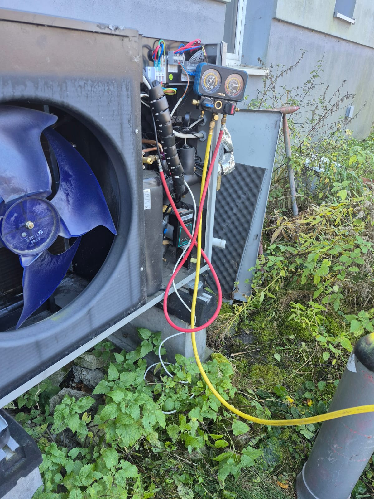 Otwarta obudowa pompy ciepła z widocznym wentylatorem i podłączonymi przewodami manometru serwisowego. Otoczenie porośnięte roślinnością.