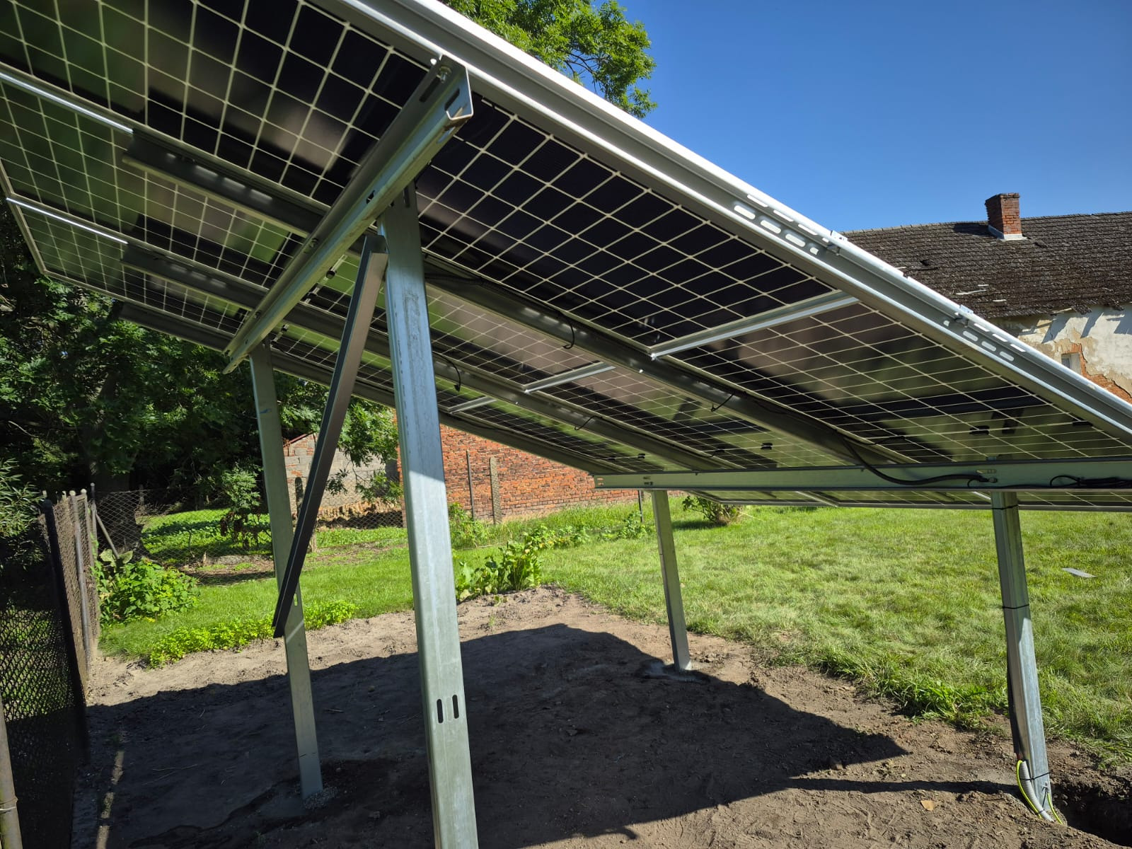 Montaż paneli fotowoltaicznych na metalowej konstrukcji w ogrodzie, widok od spodu, z fragmentem budynku w tle i błękitnym niebem.