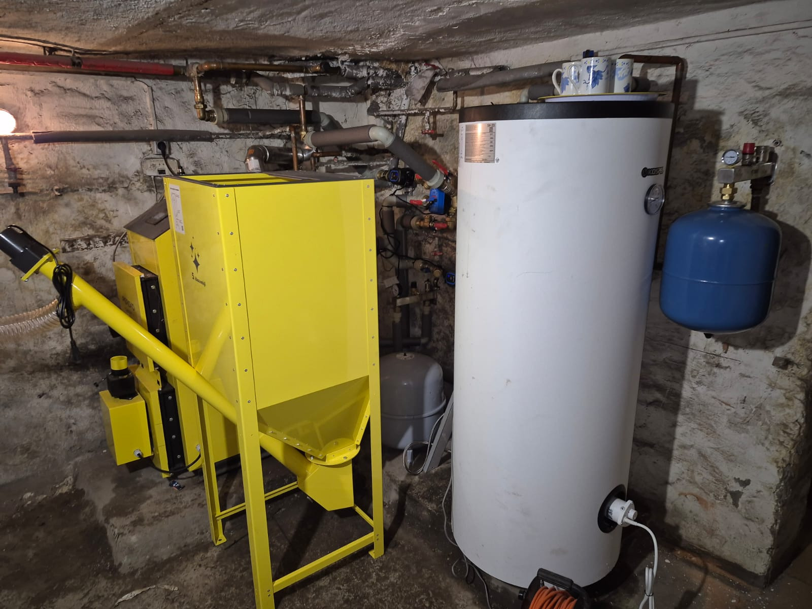 Żółty podajnik kotła na paliwo stałe obok białego zasobnika ciepłej wody w piwnicy z instalacją rurową na ścianie. Na zasobniku stoją filiżanki.