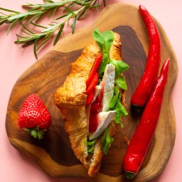 P.H.U KACPER LINKIEWICZ - Kreatywna kompozycja: croissant z serem brie, truskawkami, sałatą i sosem na drewnianej desce, obok papryczki chili i gałązka rozmarynu na różowym tle.