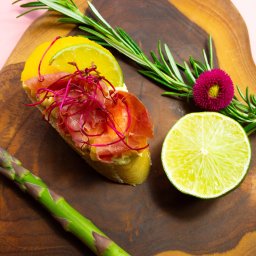 P.H.U KACPER LINKIEWICZ - Elegancka kanapka z łososiem, cytryną, limonką i rozmarynem na drewnianej desce. Całość na różowym tle, tworzy apetyczną kompozycję.