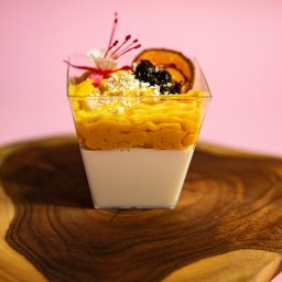 P.H.U KACPER LINKIEWICZ - Deser w przezroczystym kubeczku na drewnianej desce. Kremowe dno, żółta warstwa i dekoracja z owoców, płatków i kwiatka. Różowe tło.