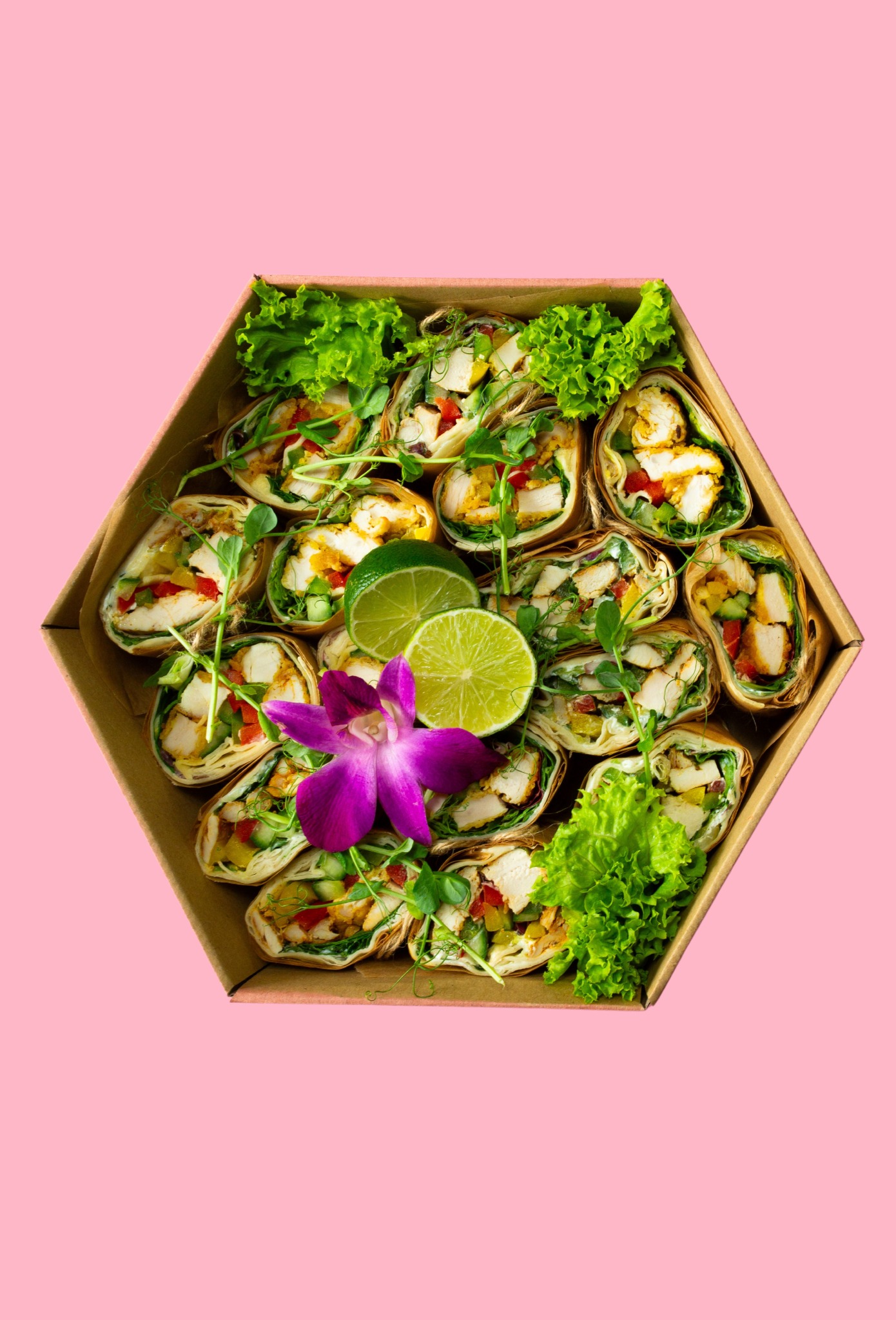 Elegancko zapakowane tortille z kurczakiem, warzywami i limonką w heksagonalnym pudełku, ozdobione kwiatem orchidei i zieleniną na różowym tle.