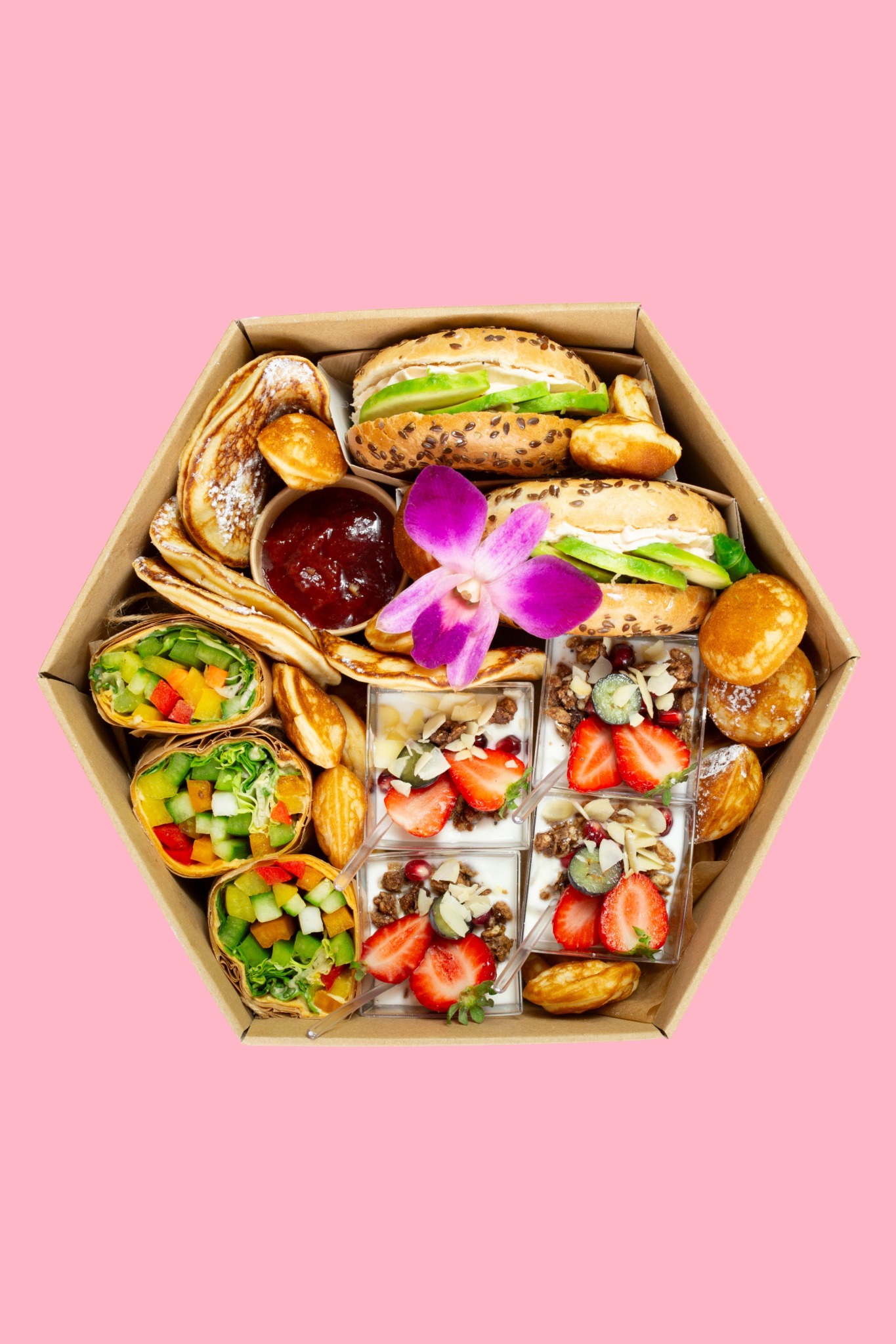 Elegancki catering w heksagonalnym pudełku: kanapki, naleśniki, wrapy warzywne i desery jogurtowe z owocami, ozdobione fioletowym kwiatem na różowym tle.