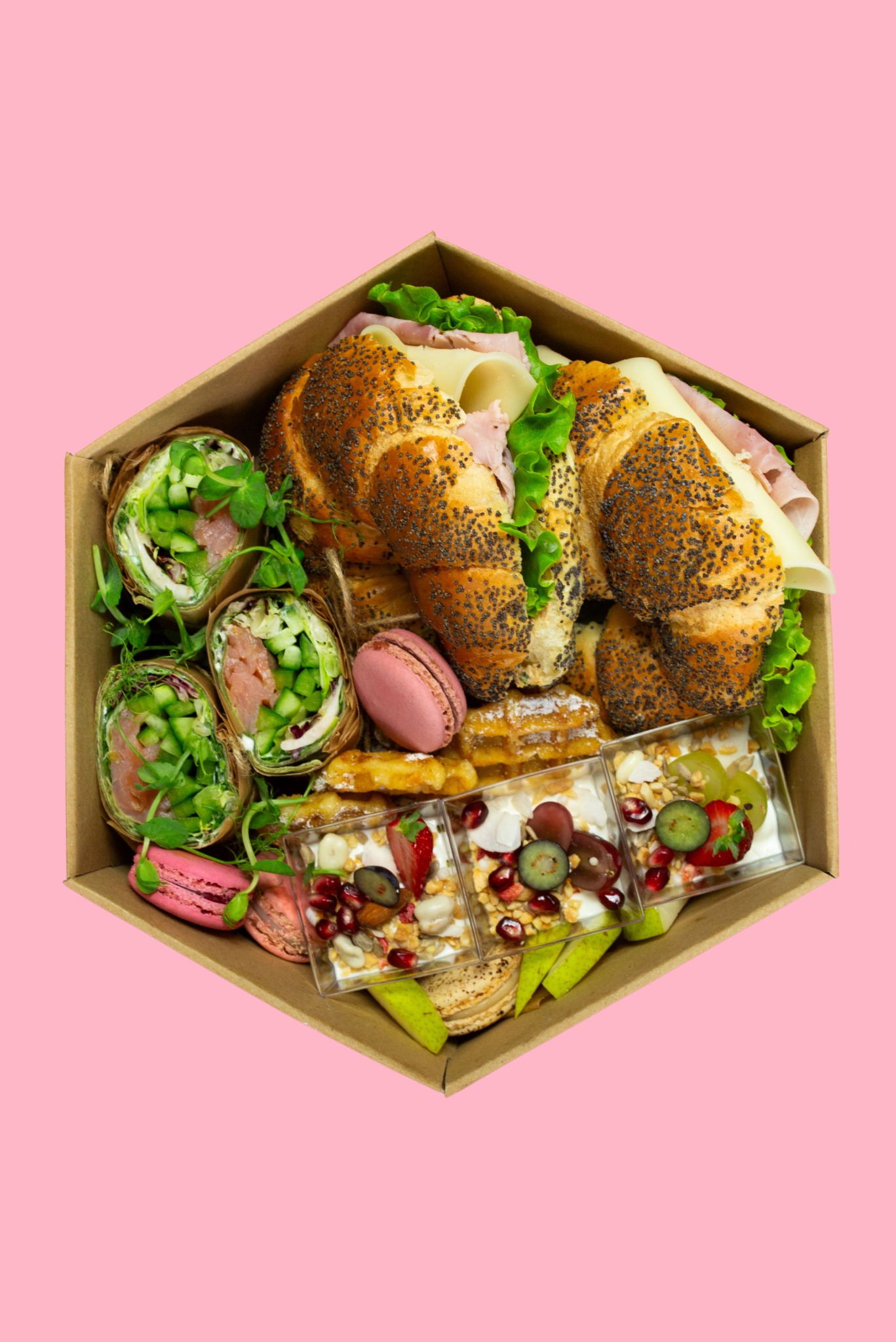 Elegancki catering w pudełku: kanapki z szynką i serem, wrapy z łososiem, mini desery z owocami i granatem, makaroniki. Całość na różowym tle.