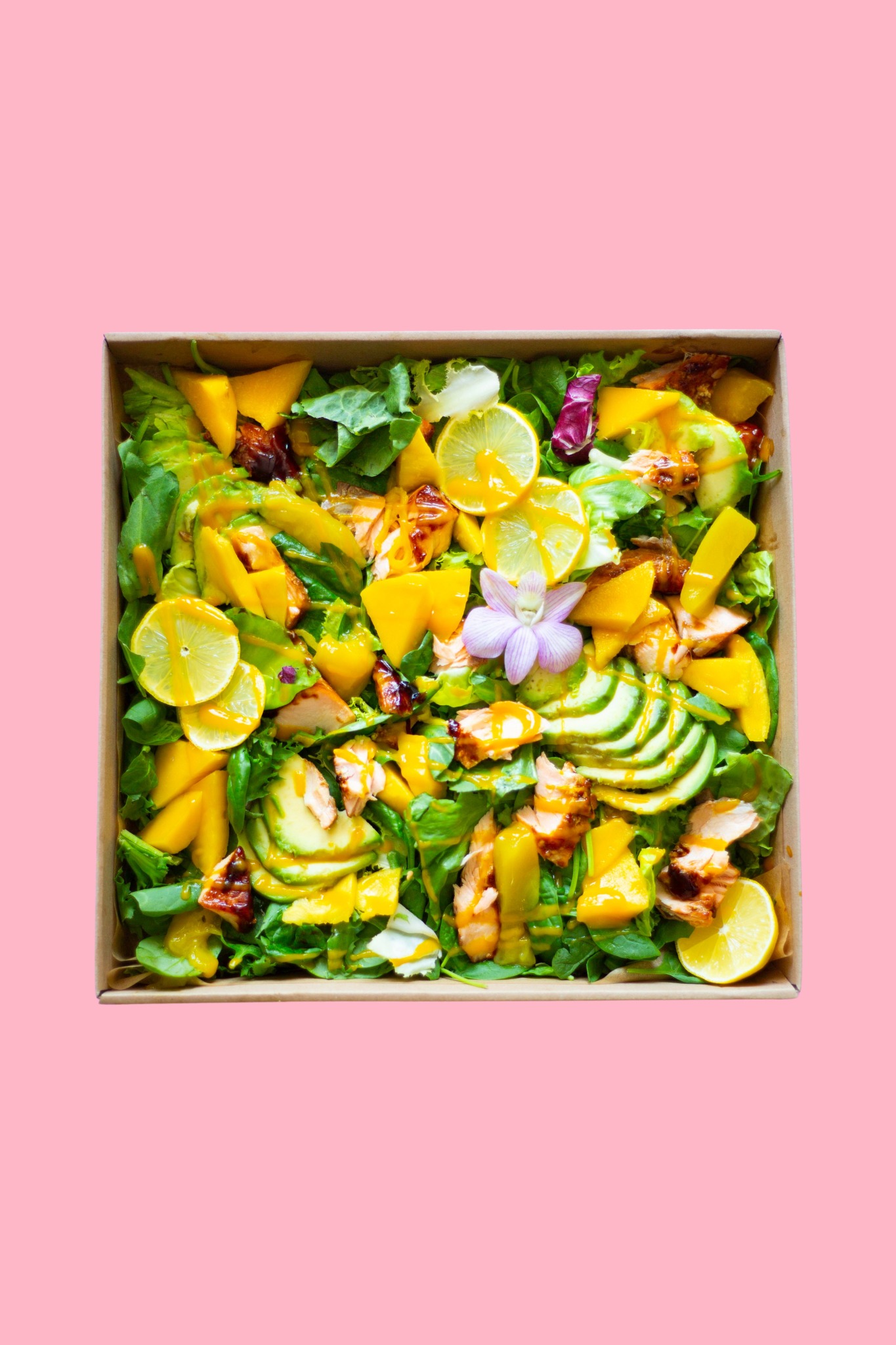 Sałatka z grillowanym kurczakiem, mango, awokado i cytryną w kartonowym pudełku na różowym tle. Dekoracja z kwiatów.