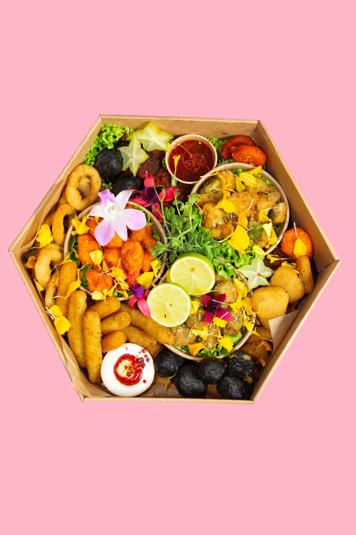 Dekoracyjna, sześciokątna patera z przekąskami: smażone warzywa, owoce, sosy, kwiaty. Całość na różowym tle, widok z góry.