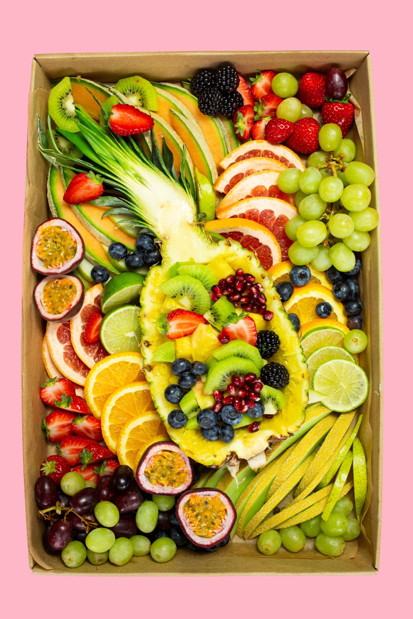 Owoce egzotyczne w kartonowym pudełku: ananas, kiwi, marakuja, melon, pomarańcze, grejpfruty, truskawki, jeżyny, borówki i winogrona na różowym tle.