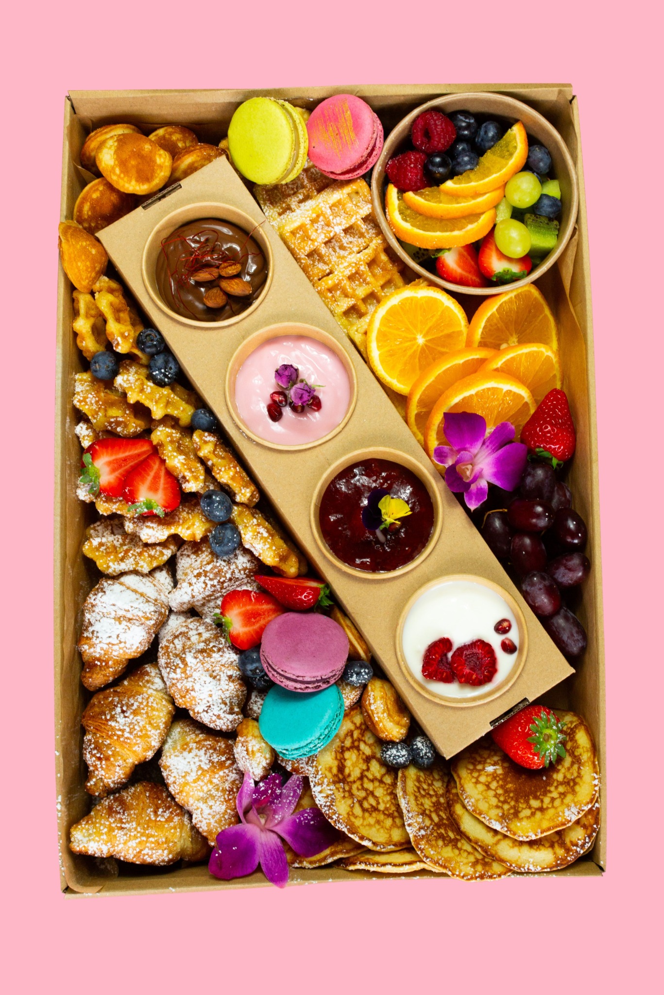 Elegancki box śniadaniowy: mini naleśniki, gofry, croissanty z cukrem pudrem, owoce, makaroniki i sosy w papierowych miseczkach, całość na różowym tle.