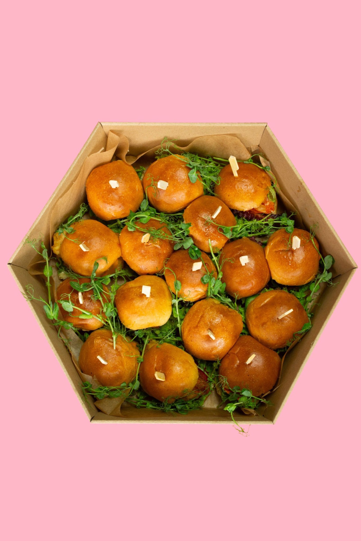Zestaw mini burgerów w kartonowym pudełku, udekorowany zielonymi pędami, na różowym tle. Burgery z jasną bułką i małym kawałkiem sera na wierzchu, idealne na catering.