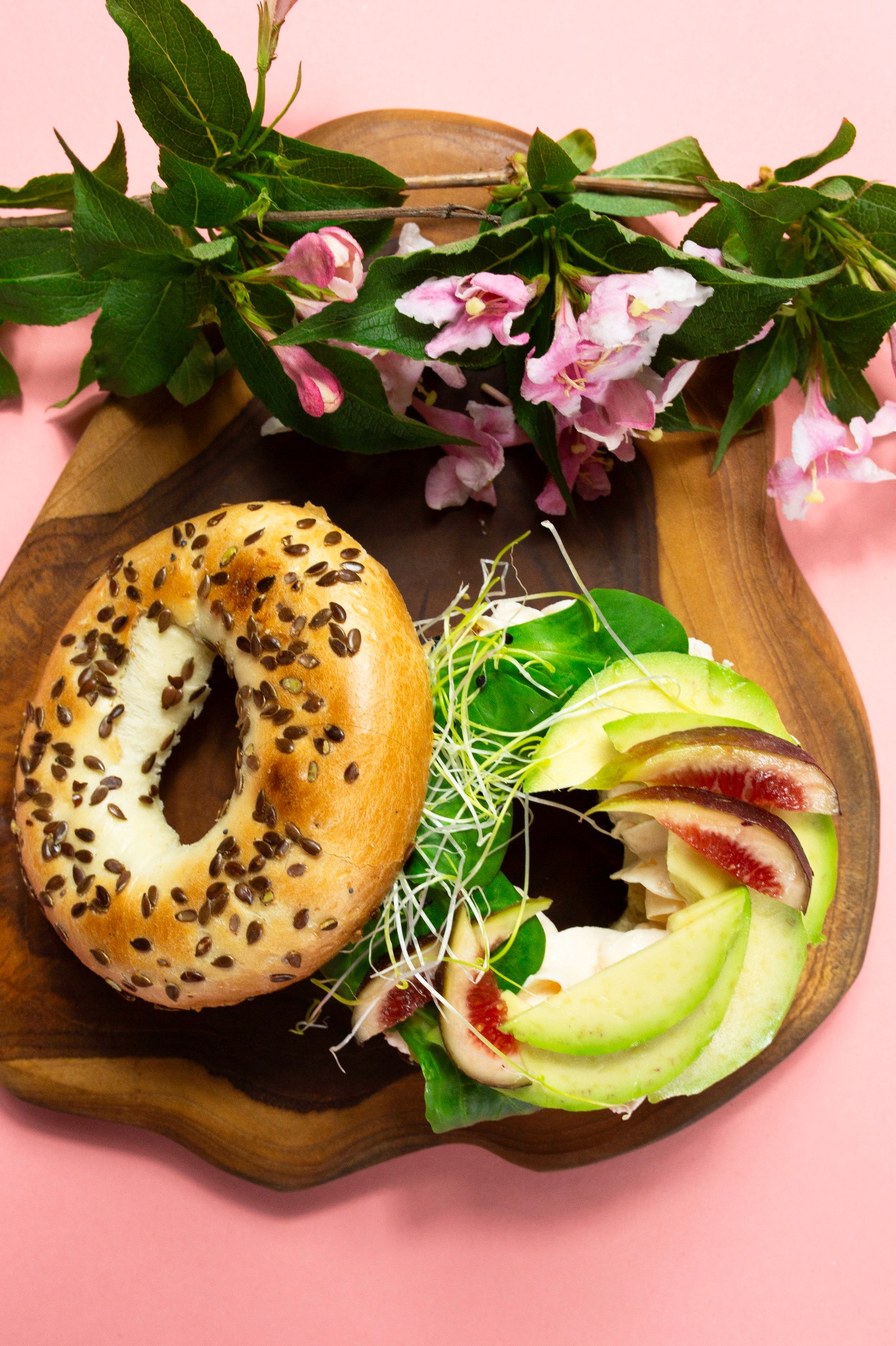 Bagel z awokado, figami i kiełkami na drewnianej desce, ozdobiony gałązką kwitnącej wiśni na różowym tle. Zdjęcie z góry, jasne oświetlenie.