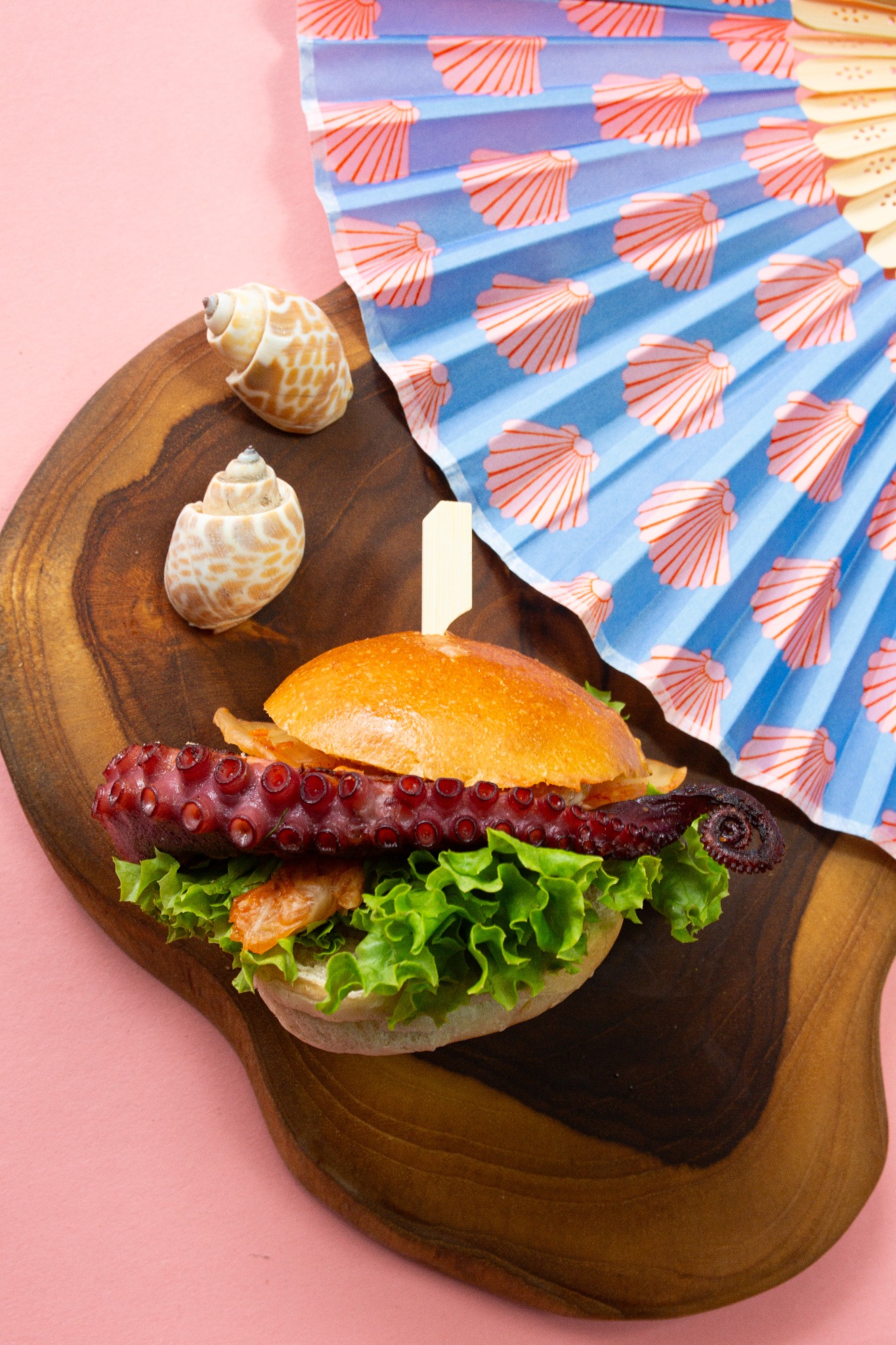 Burger z ośmiornicą na drewnianej desce, muszle i wachlarz z motywem muszelek w tle. Nietypowa kompozycja kulinarna na różowym tle.
