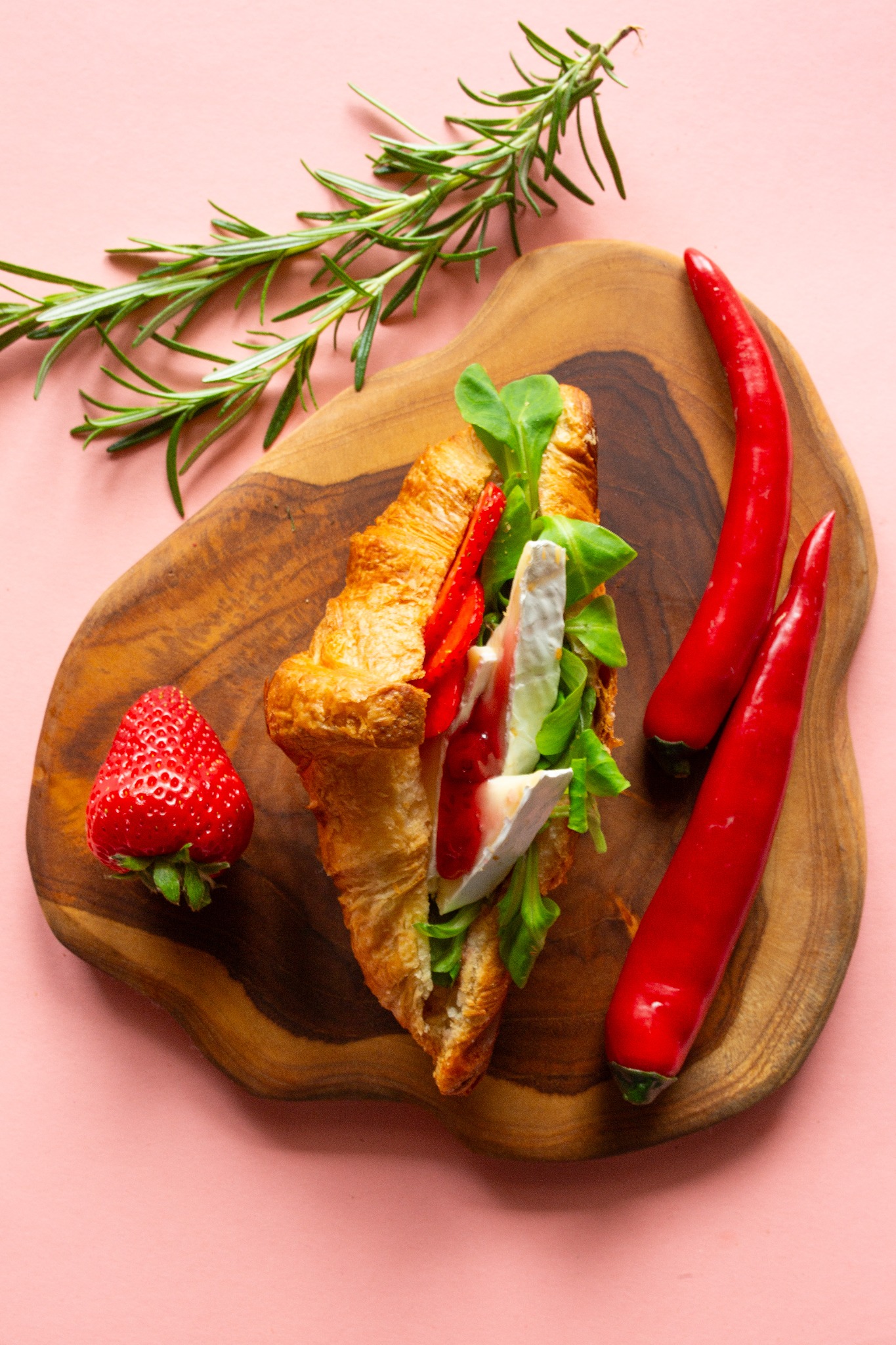 Kreatywna kompozycja: croissant z serem brie, truskawkami, sałatą i sosem na drewnianej desce, obok papryczki chili i gałązka rozmarynu na różowym tle.