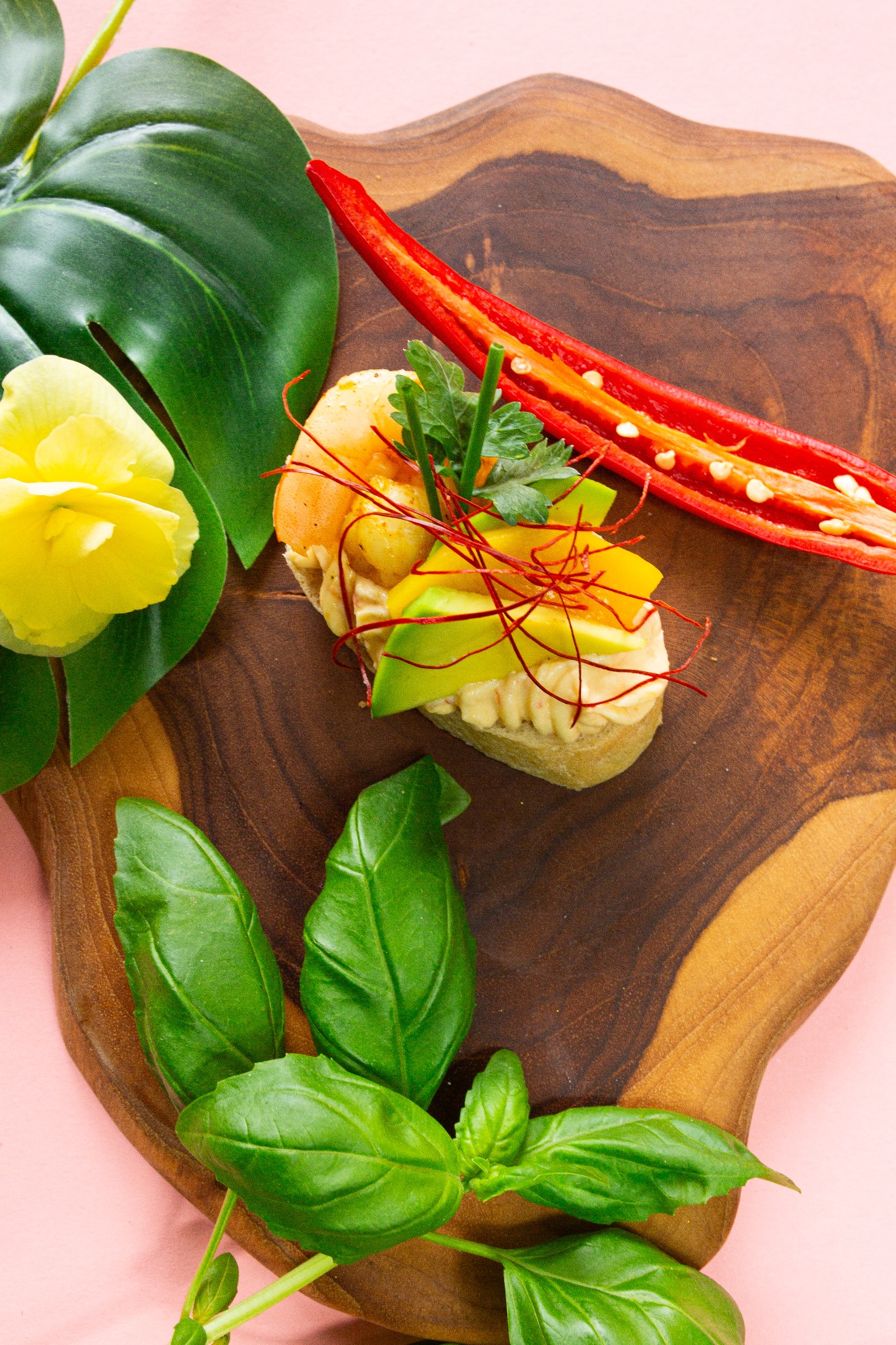 Apetyczna kanapka z krewetką, awokado i chili na drewnianej desce, ozdobiona bazylią, żółtym kwiatem i liściem monstery. Jasnoróżowe tło. Elegancka kompozycja.