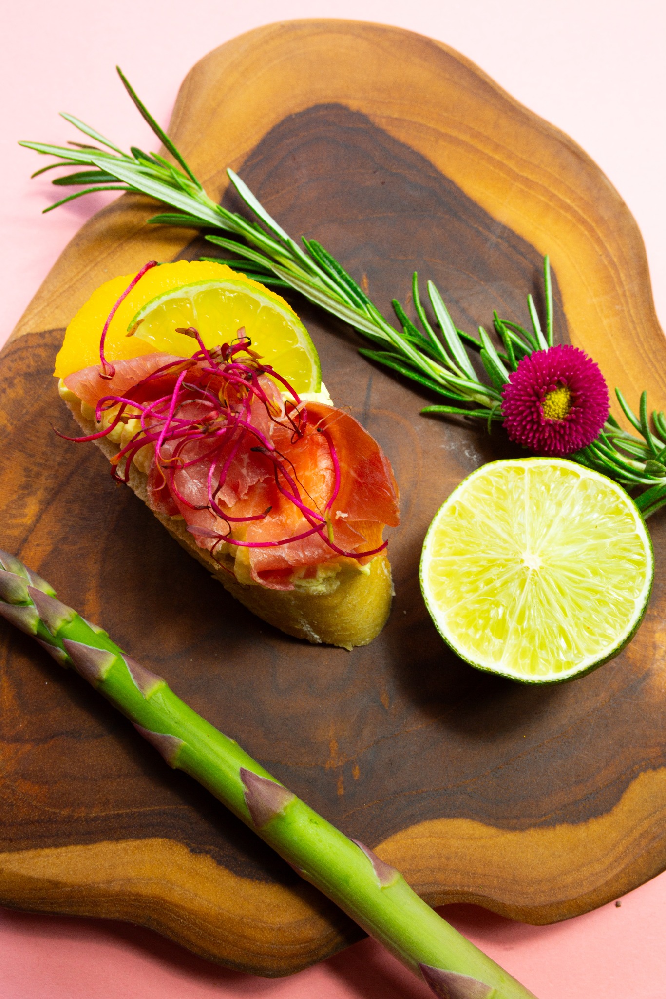Elegancka kanapka z łososiem, cytryną, limonką i rozmarynem na drewnianej desce. Całość na różowym tle, tworzy apetyczną kompozycję.