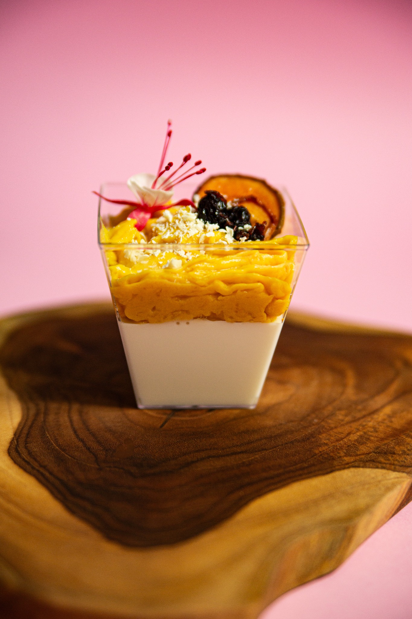 Deser w przezroczystym kubeczku na drewnianej desce. Kremowe dno, żółta warstwa i dekoracja z owoców, płatków i kwiatka. Różowe tło.