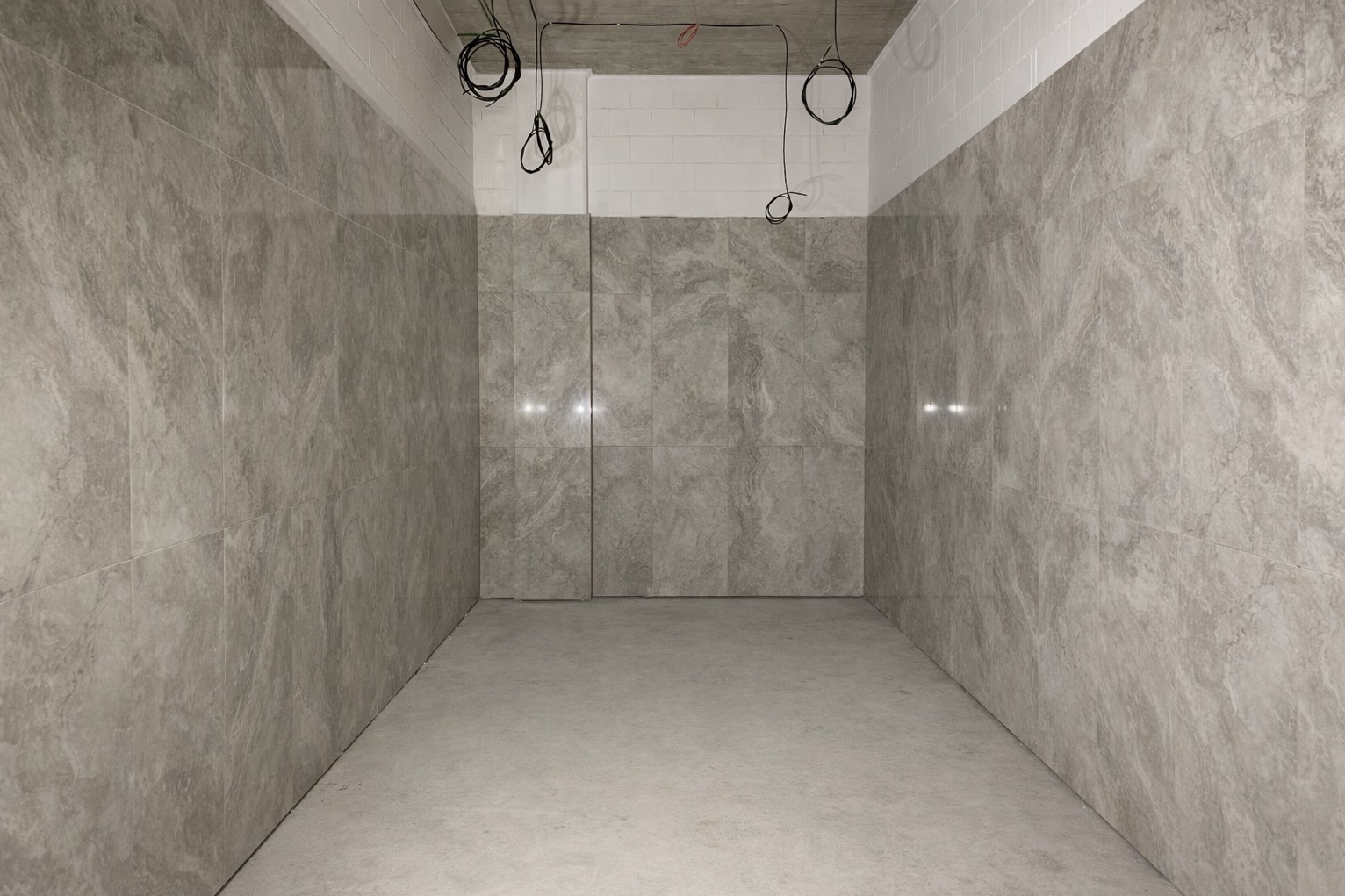 Wykończona łazienka z szarymi, marmurkowymi płytkami na ścianach i podłodze. Widoczne przewody elektryczne na suficie. Minimalistyczny design, surowy betonowy sufit.