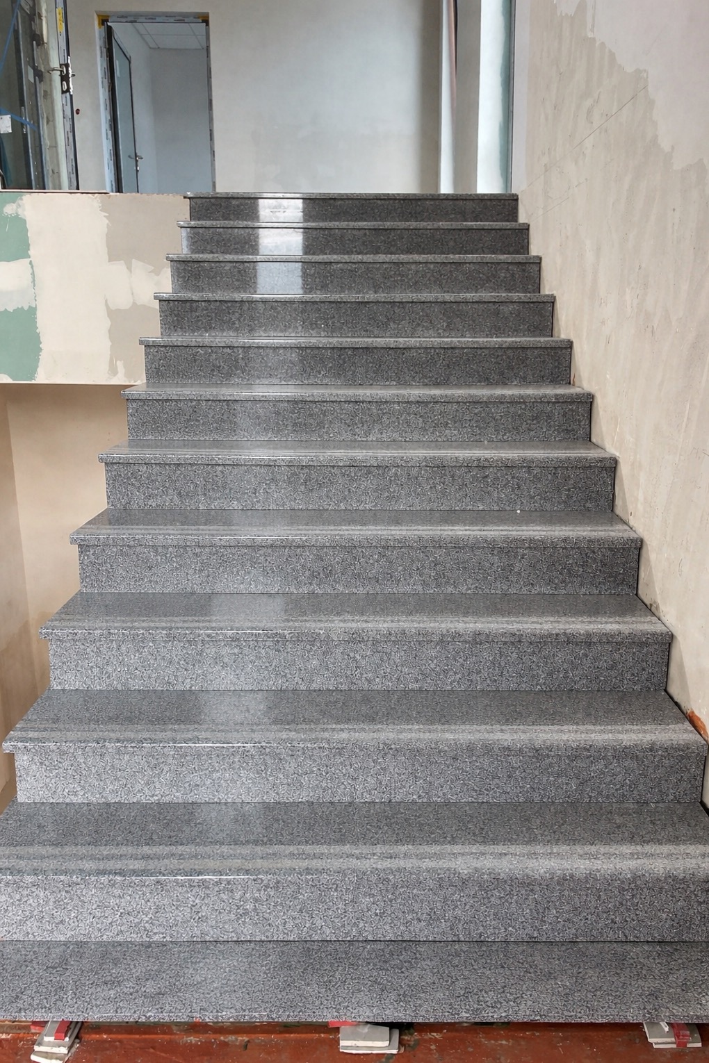 Granitowe schody w trakcie wykończenia, widoczne niedoskonałości ścian i drzwi w tle. Ujęcie z dołu, podkreślające strukturę kamienia i etap prac wykończeniowych.