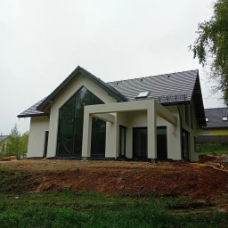 Nowoczesny dom jednorodzinny z białymi ścianami i dużymi oknami, częściowo zakryty folią ochronną, na etapie wykończenia elewacji. Widoczne elementy konstrukcyjne.