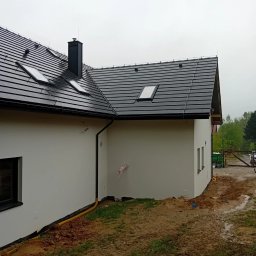 Biała elewacja domu jednorodzinnego z czarną dachówką i oknami dachowymi, widok z boku na tle pochmurnego nieba i błotnistego terenu.