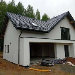 Biały dom jednorodzinny z garażem w trakcie wykończenia elewacji, widoczne rury i materiały budowlane na zewnątrz. Dach pokryty ciemną dachówką.