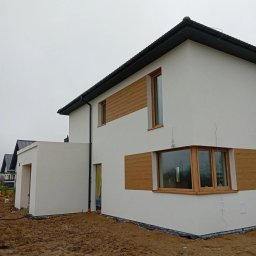 Nowoczesny dom z białą elewacją i drewnianymi akcentami wokół okien. Widoczne detale wykończenia i otoczenie domu w trakcie prac ziemnych.