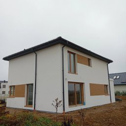 NEST-UP - Dwukondygnacyjny dom z białą elewacją i drewnianymi akcentami, widoczny z zewnątrz na tle pochmurnego nieba i przygotowanego terenu.