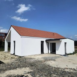 NEST-UP - Biały dom jednorodzinny z czerwoną dachówką, widoczny w słoneczny dzień. Minimalistyczna bryła, czarne ramy okienne, surowe otoczenie placu budowy.