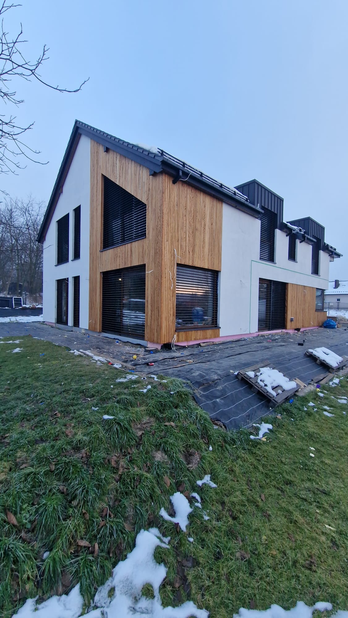 Nowoczesny dom z elementami drewnianej elewacji i dużymi oknami z żaluzjami zewnętrznymi. Widoczne prace wykończeniowe wokół budynku.